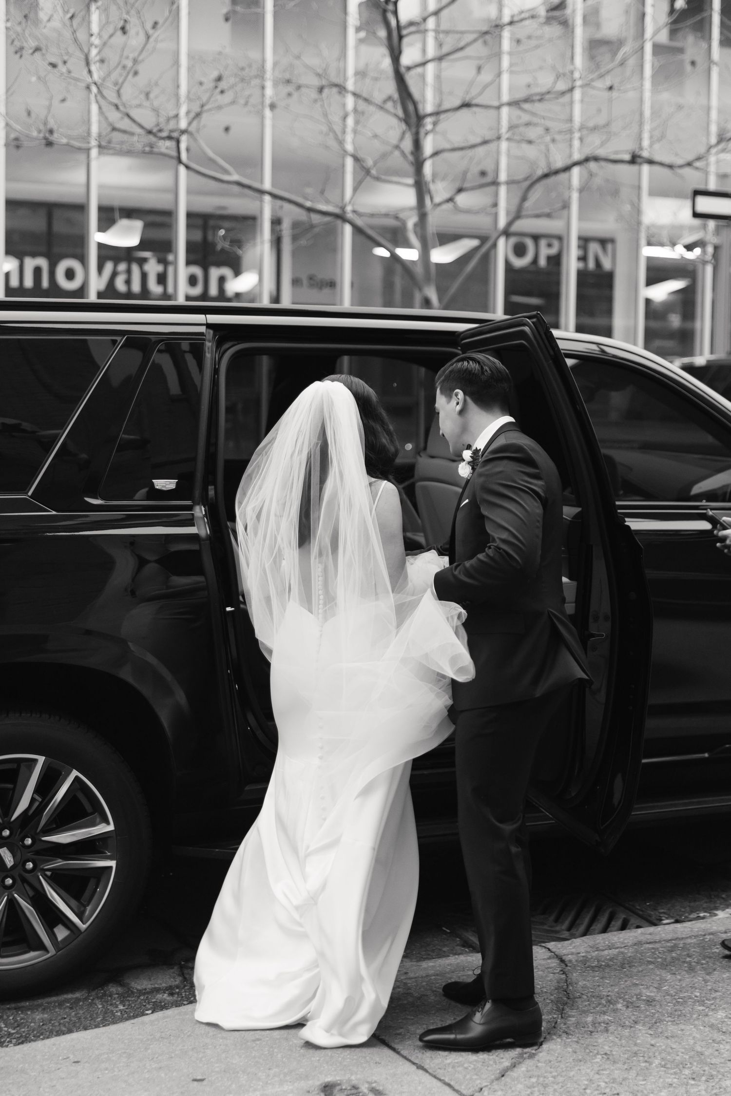 A couple in formal wedding attire preparing to enter a black SUV on a city street.
