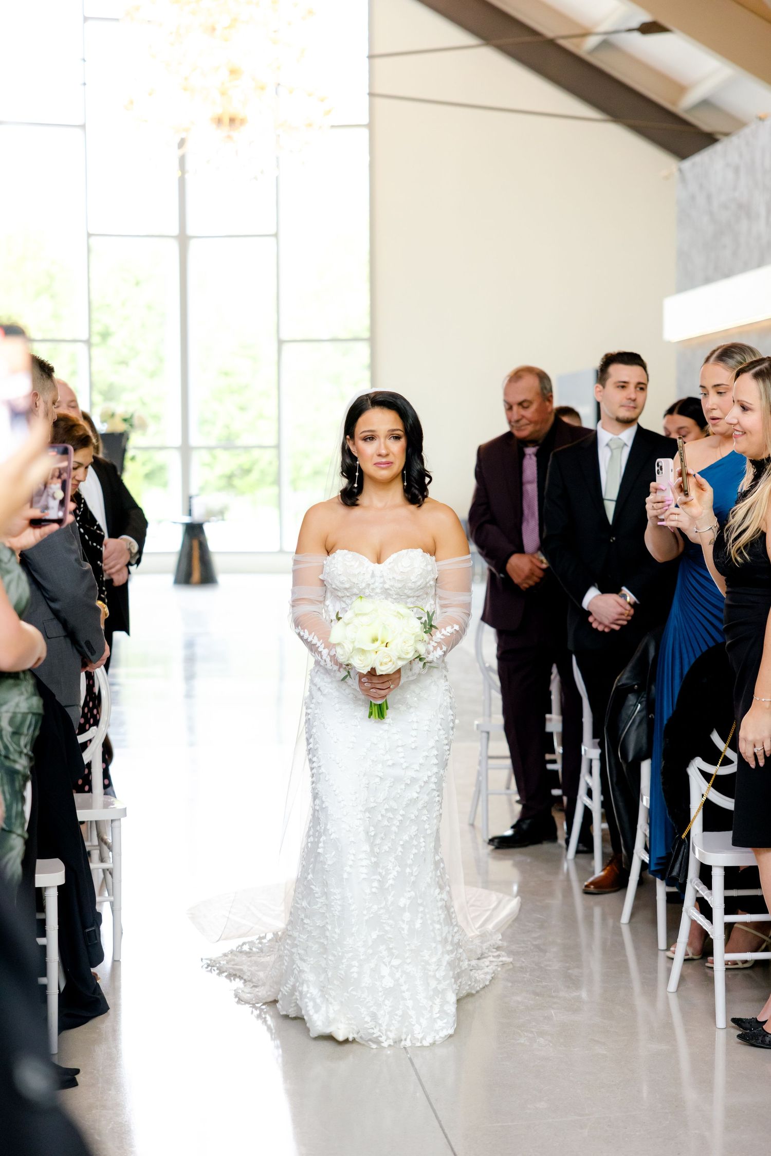 Elegant wedding ceremony in modern white venue with guests lining the aisle.