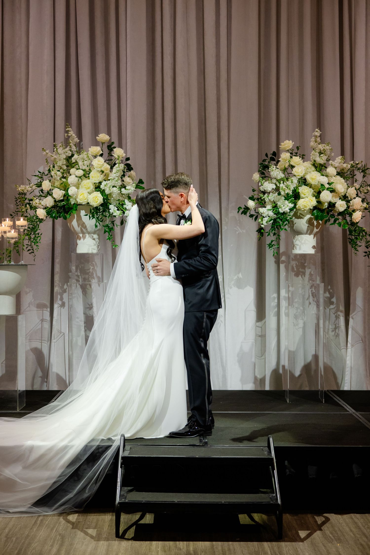 Newlyweds share intimate moment on stage between beautiful white and green floral arrangements.