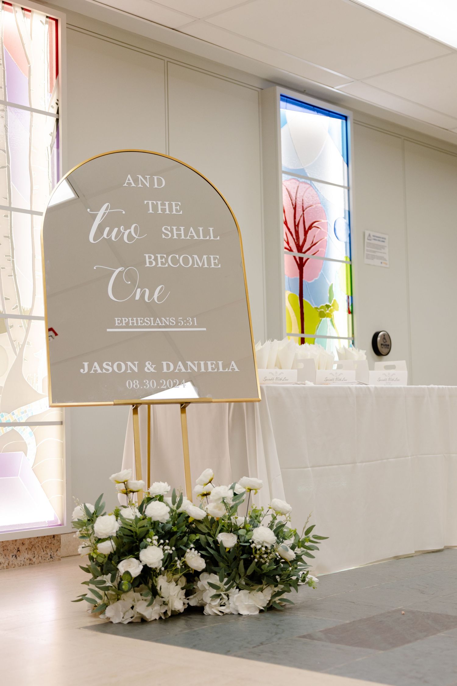 Wedding sign displays 'And the two shall become one' with elegant calligraphy and floral arrangements beneath.