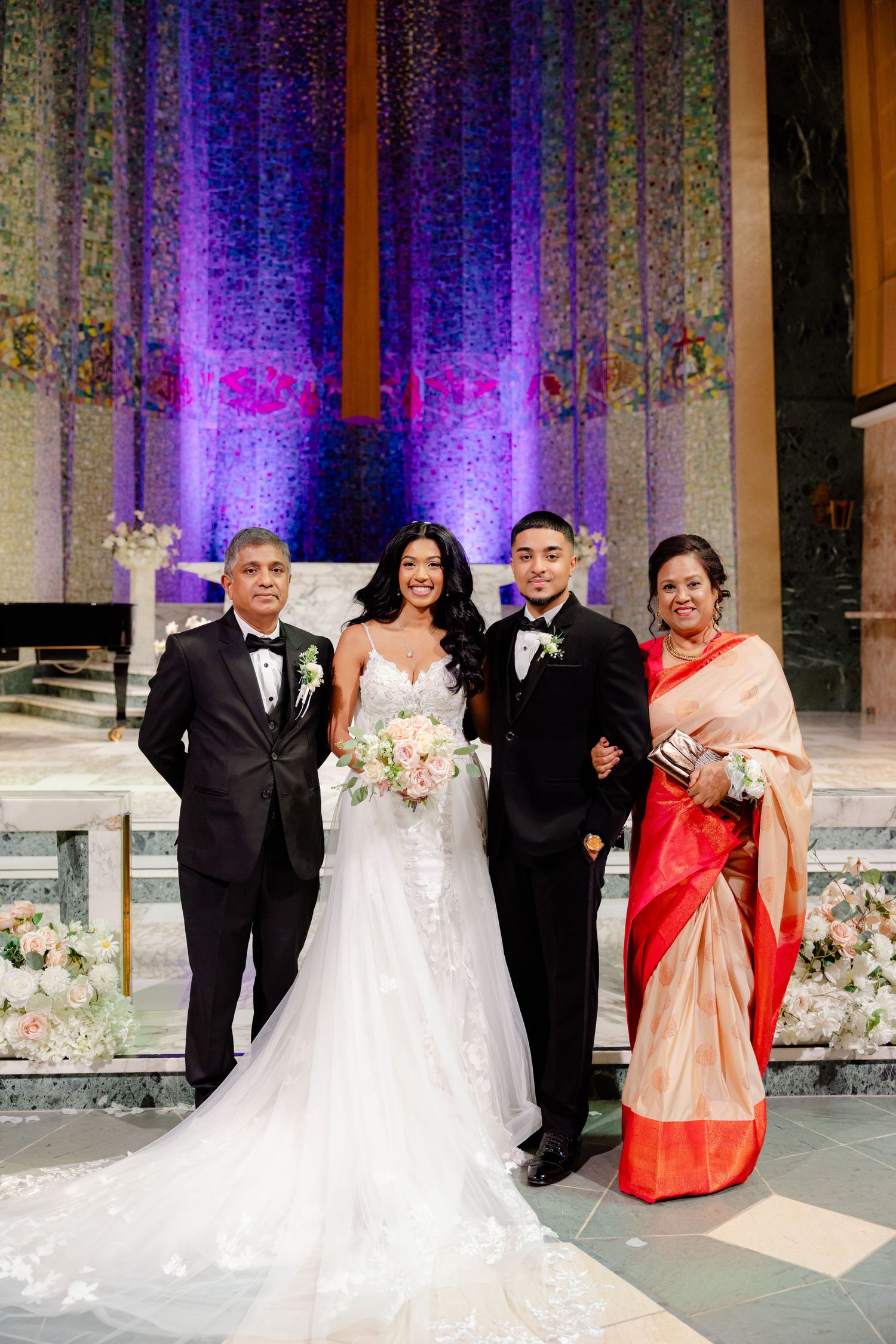 Wedding party poses at church altar illuminated by dramatic purple lighting and adorned with white flowers.