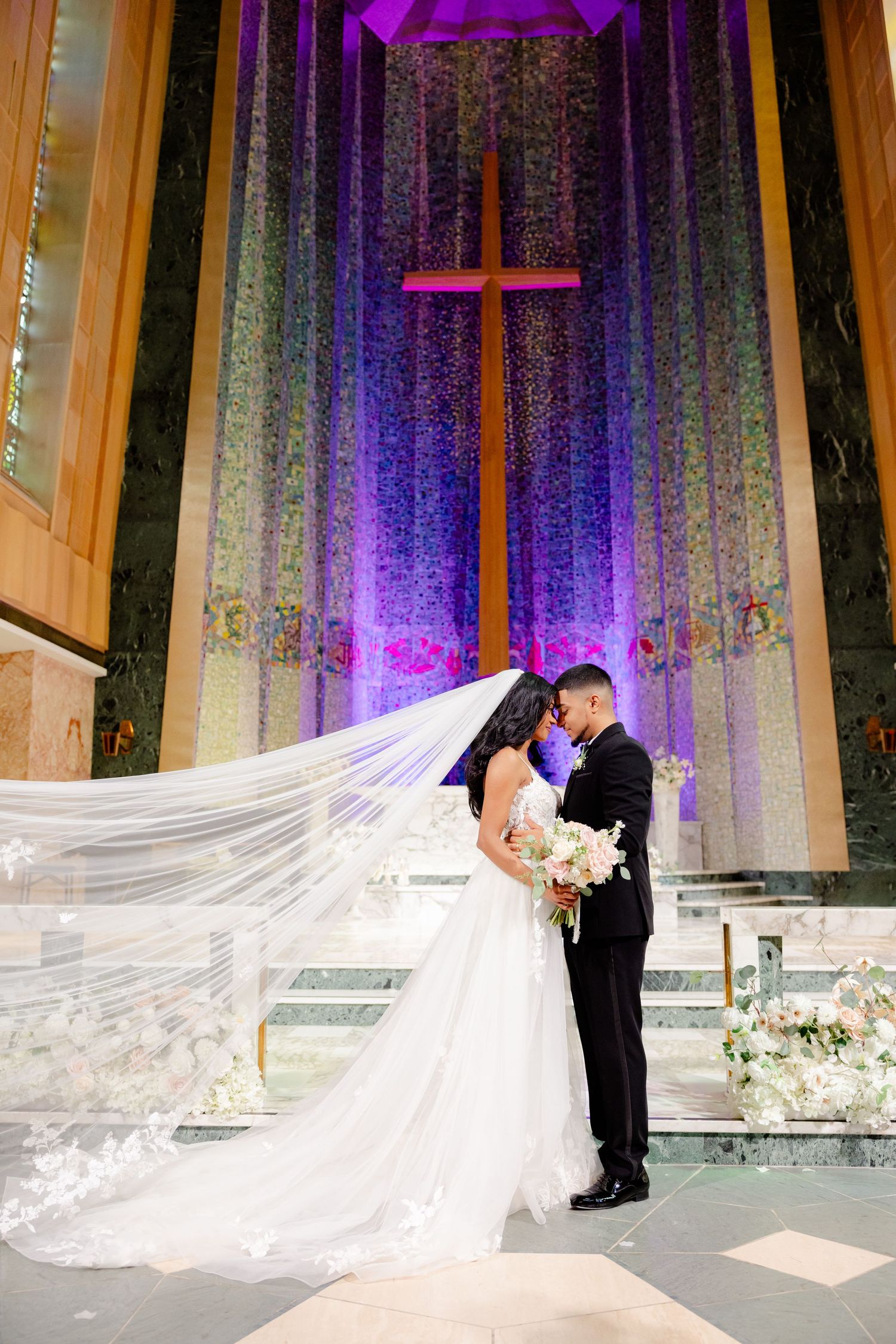 Intimate wedding kiss under dramatic purple-lit cross in modern church sanctuary.