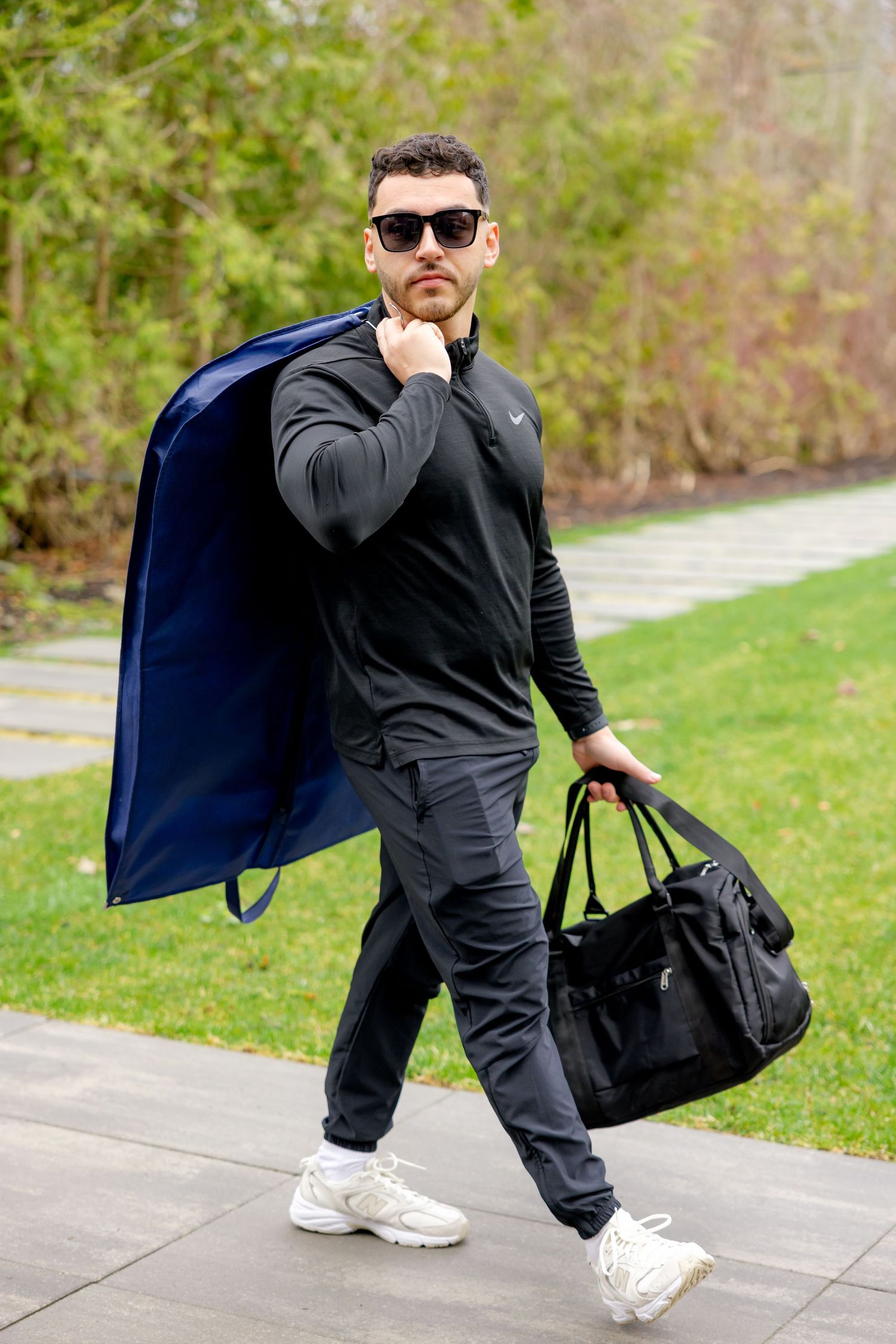 Figure in dark athletic wear carrying blue coat and black duffle bag outdoors.