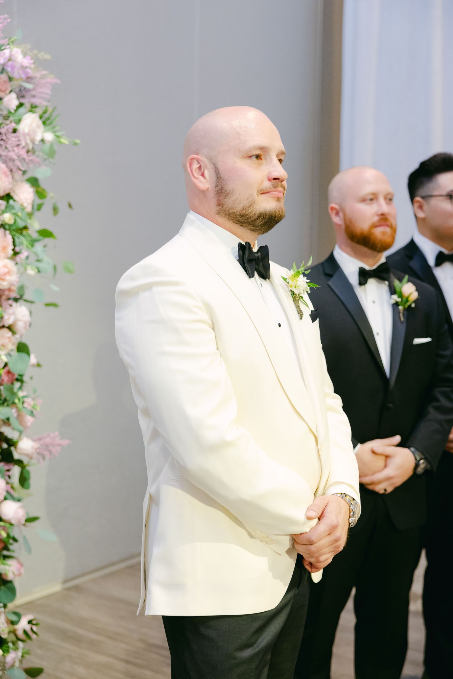 Groom and groomsmen stand at altar with pink floral arrangements during wedding ceremony.