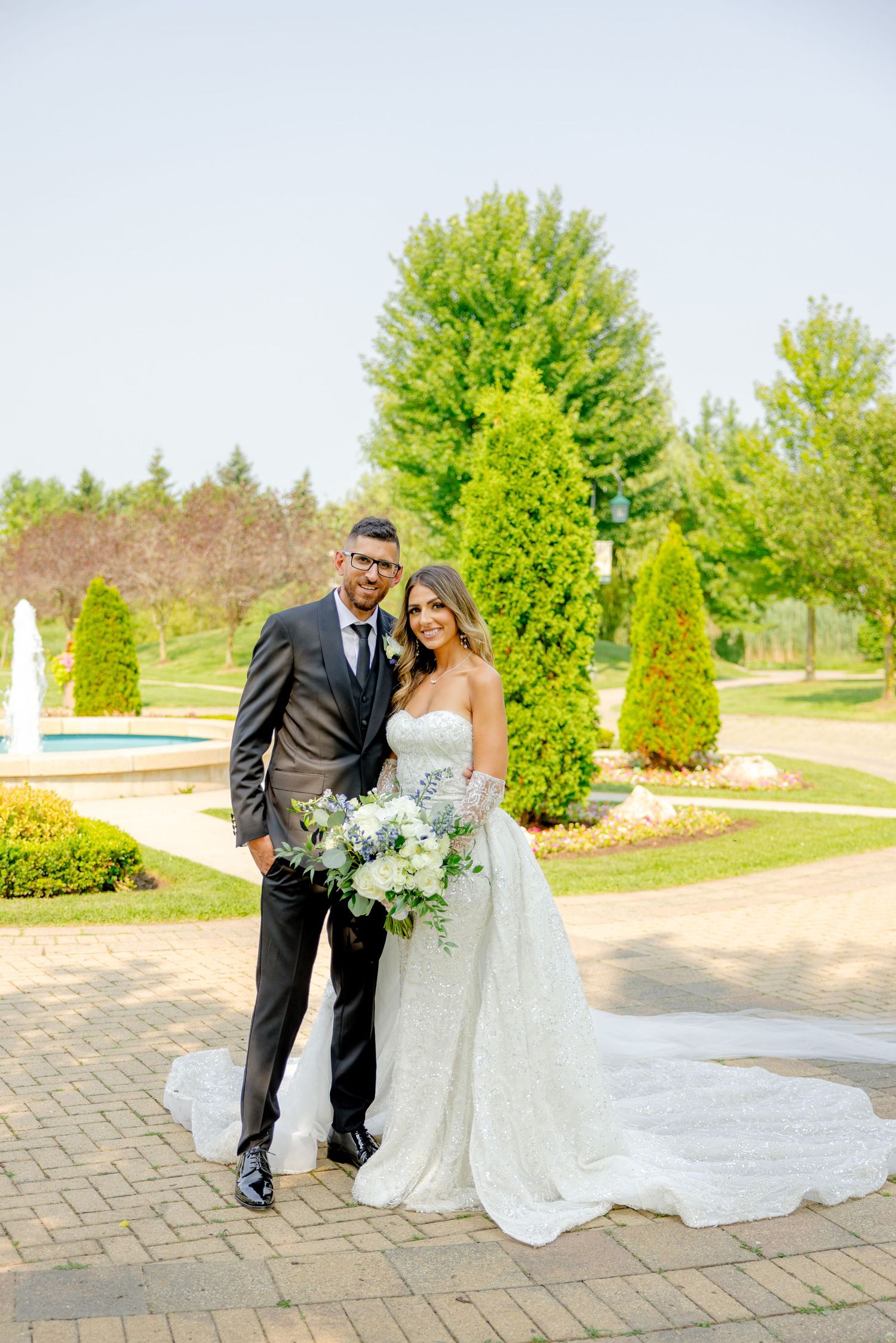 Elegant wedding couple poses on brick pathway in manicured garden with fountain.