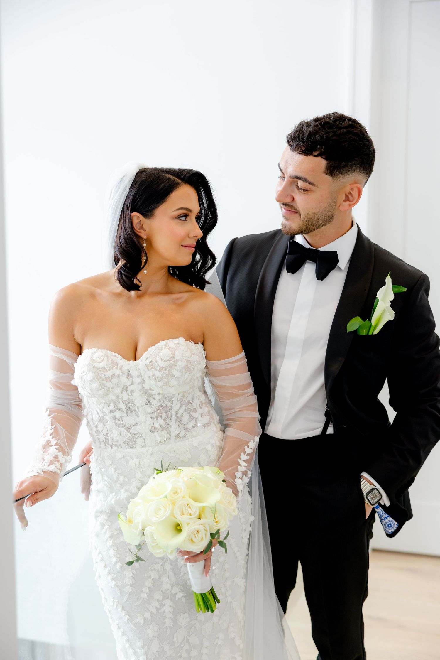 Elegant couple in wedding attire posing against white wall with bouquet.