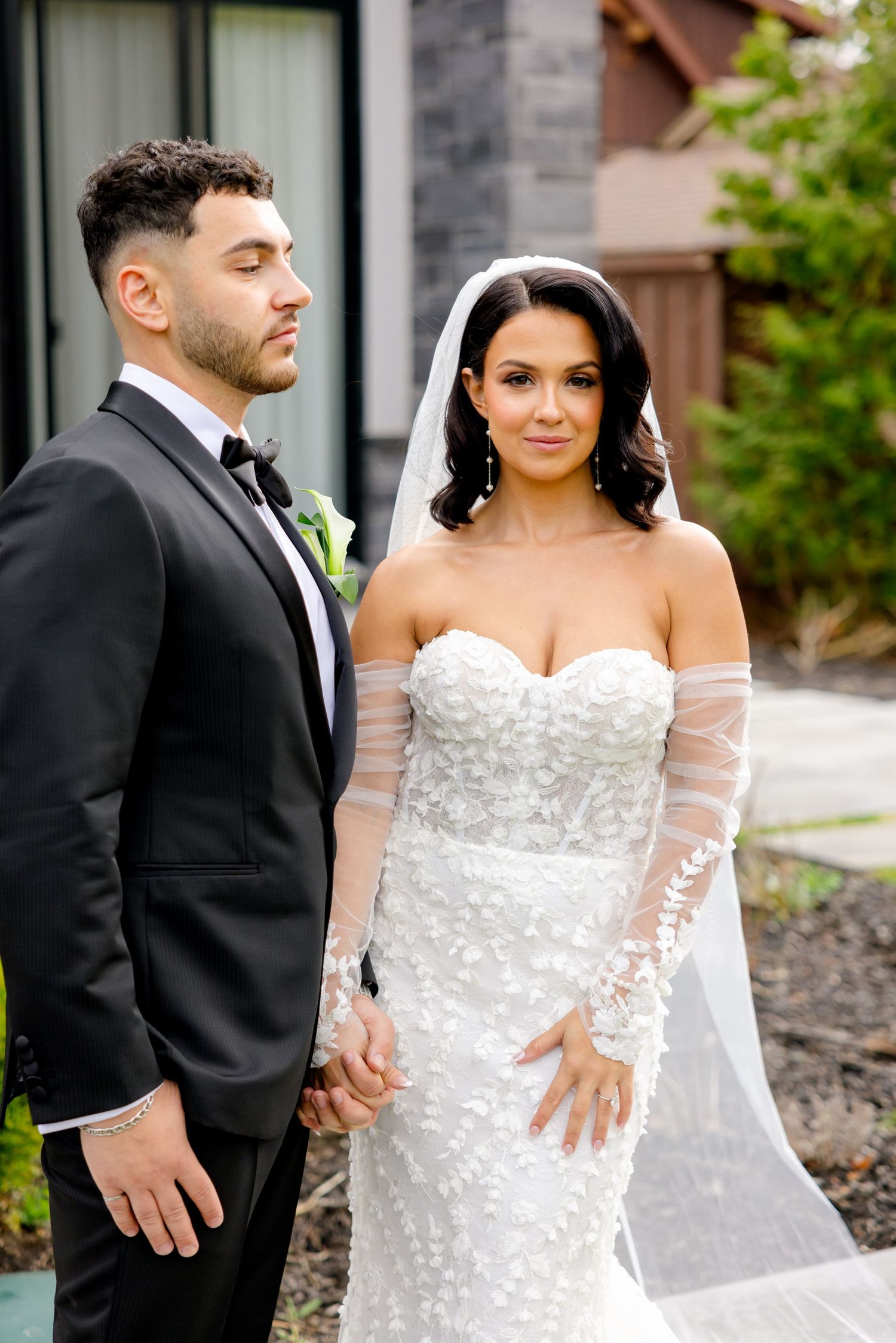 A stunning wedding portrait featuring formal attire and a detailed lace gown against stone architecture.