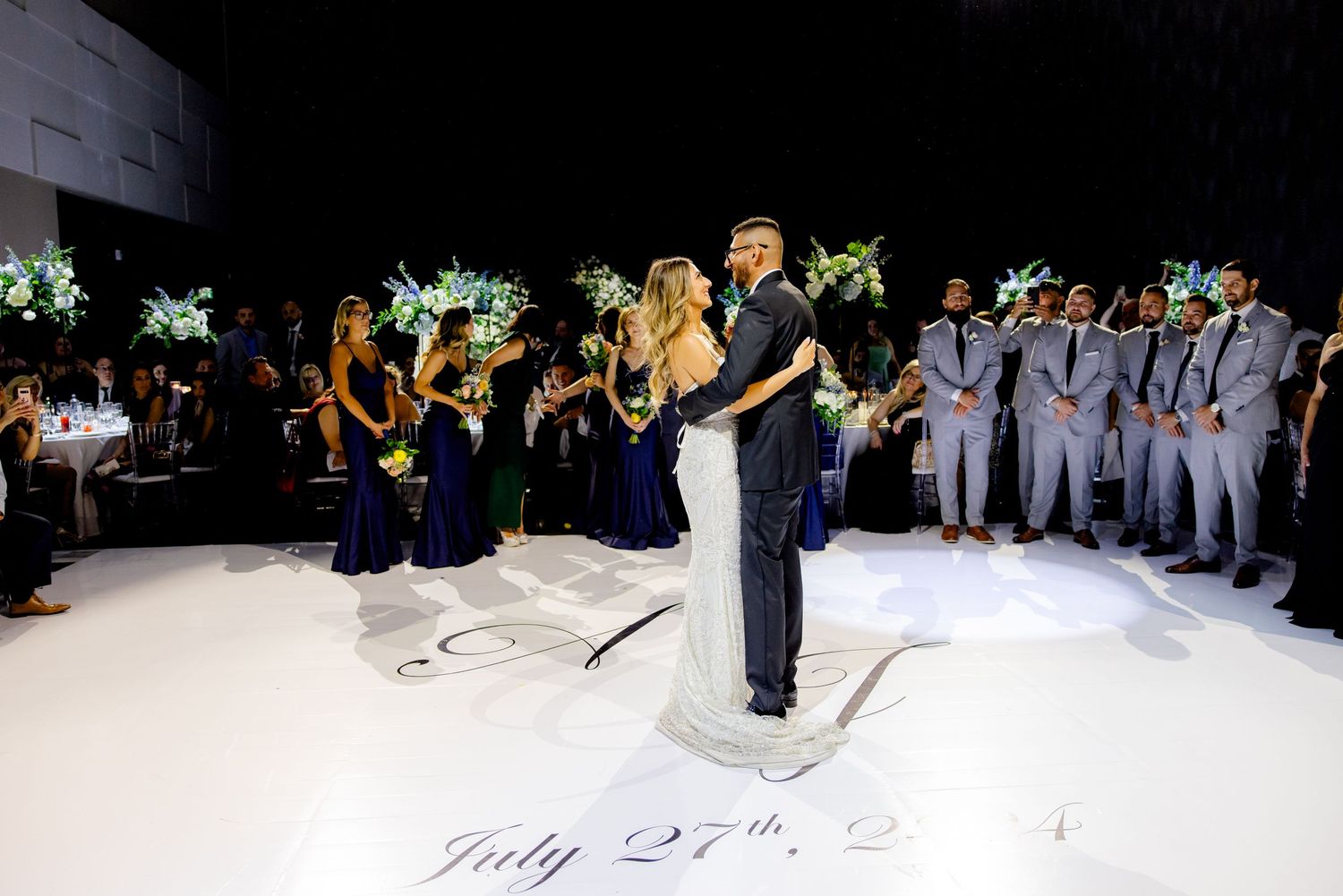 A couple dances on a white floor with date inscription while wedding party watches from both sides.