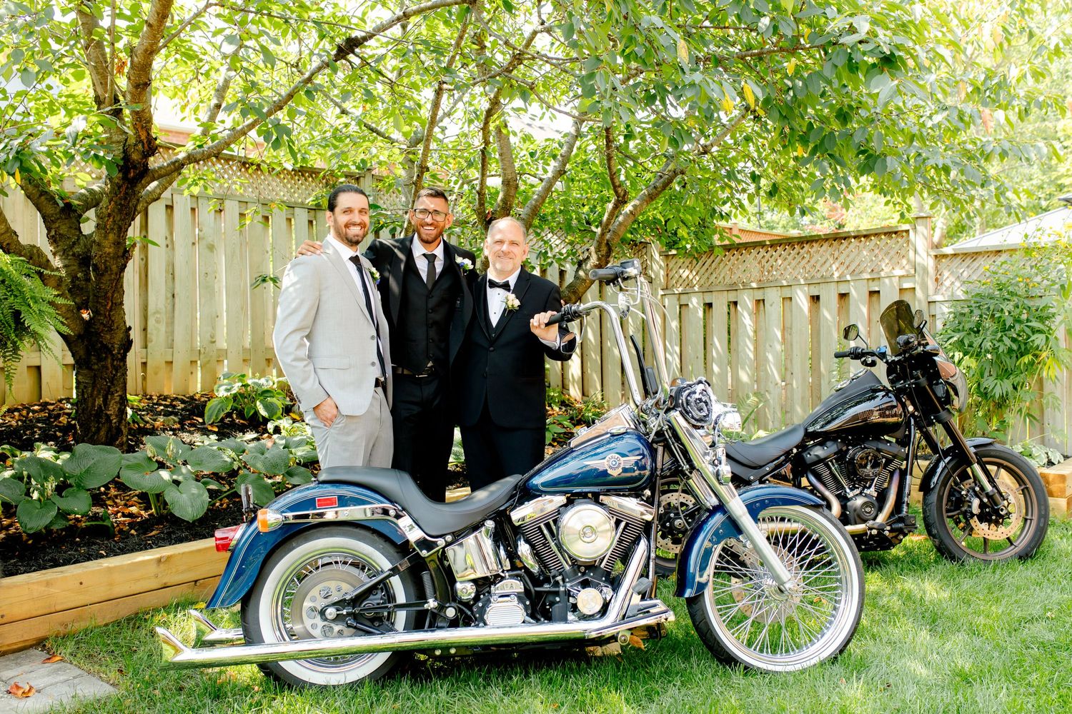 Three groomsmen pose with vintage motorcycles in a landscaped backyard setting.