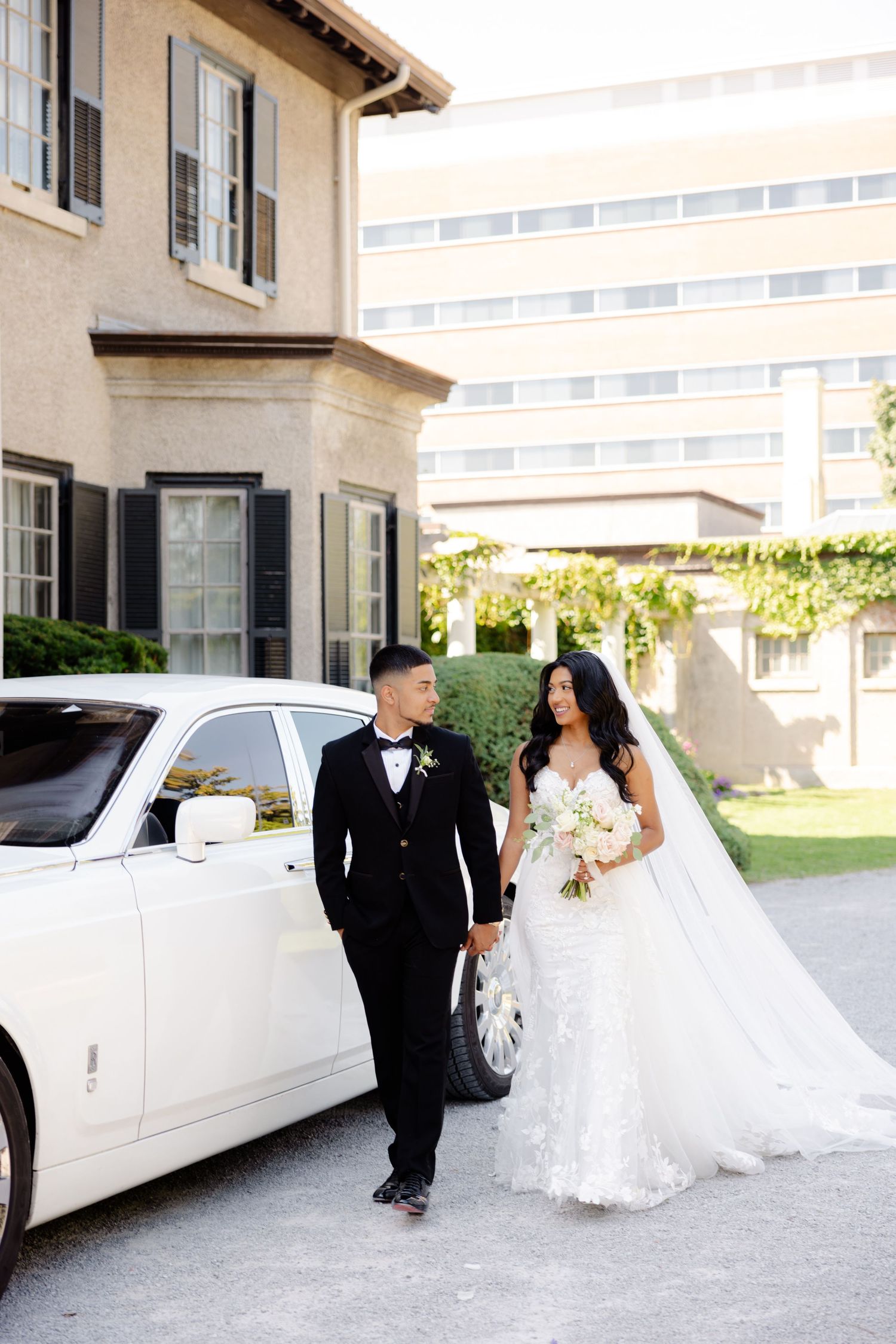 A wedding couple walking hand in hand past their white Rolls-Royce at an upscale venue.