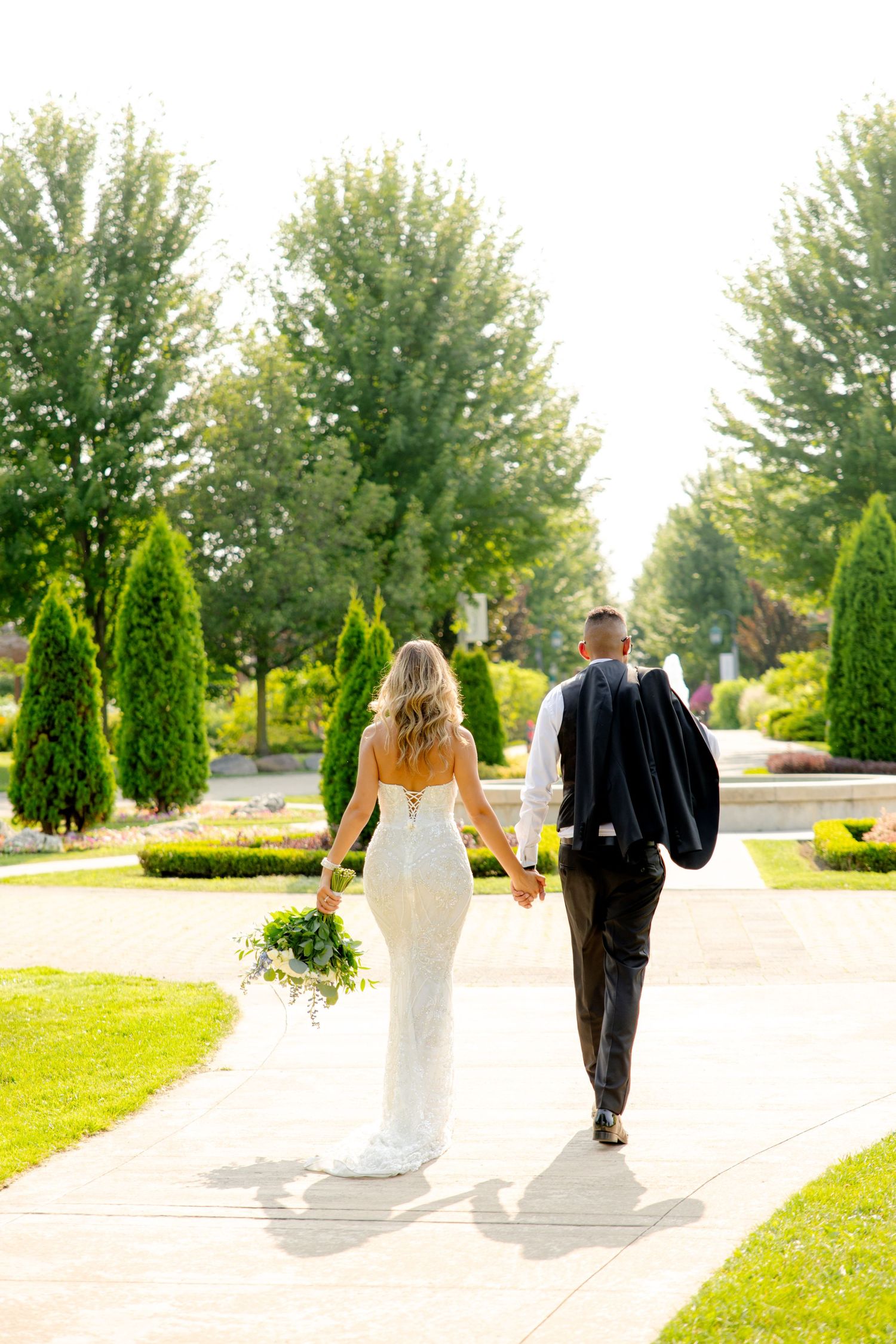 Newlyweds walking down a landscaped pathway with the bride holding a bouquet and groom carrying his jacket.