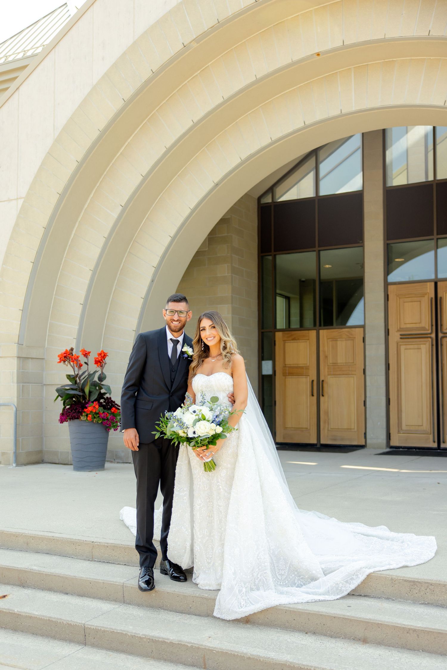 Wedding couple poses on stone steps outside modern church building with arched architecture.