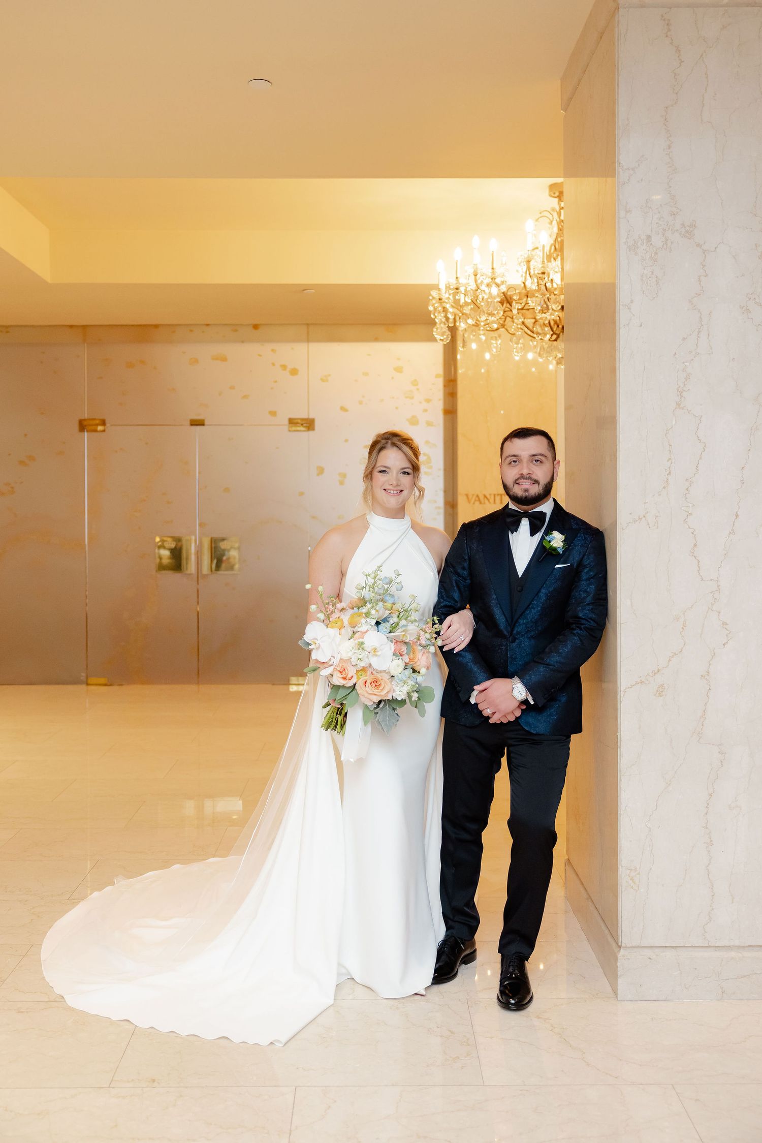 Wedding couple walks through luxurious hallway with crystal chandeliers and marble walls.