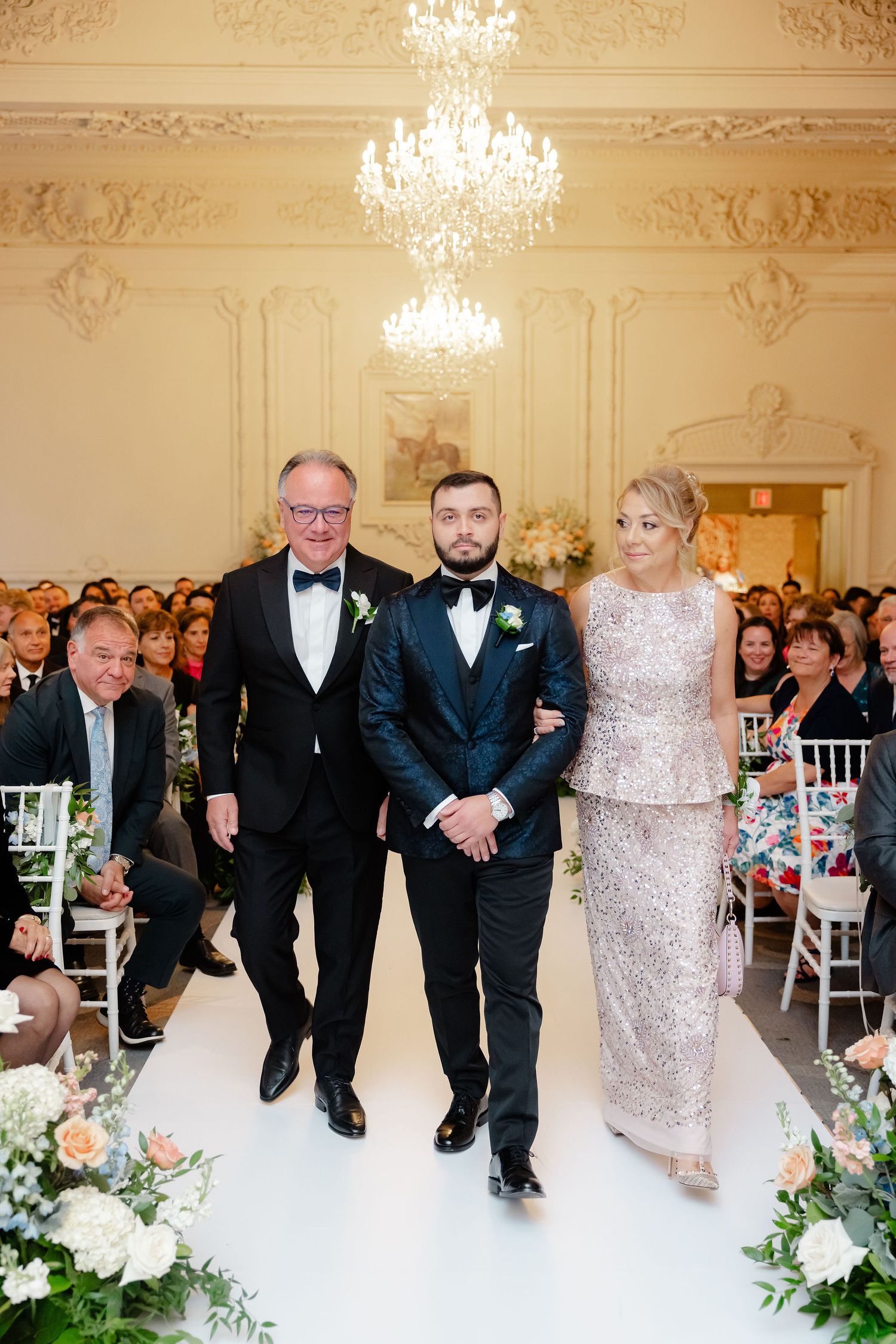 Wedding party members in formal attire walking down the aisle in an elegant ballroom with crystal chandeliers.