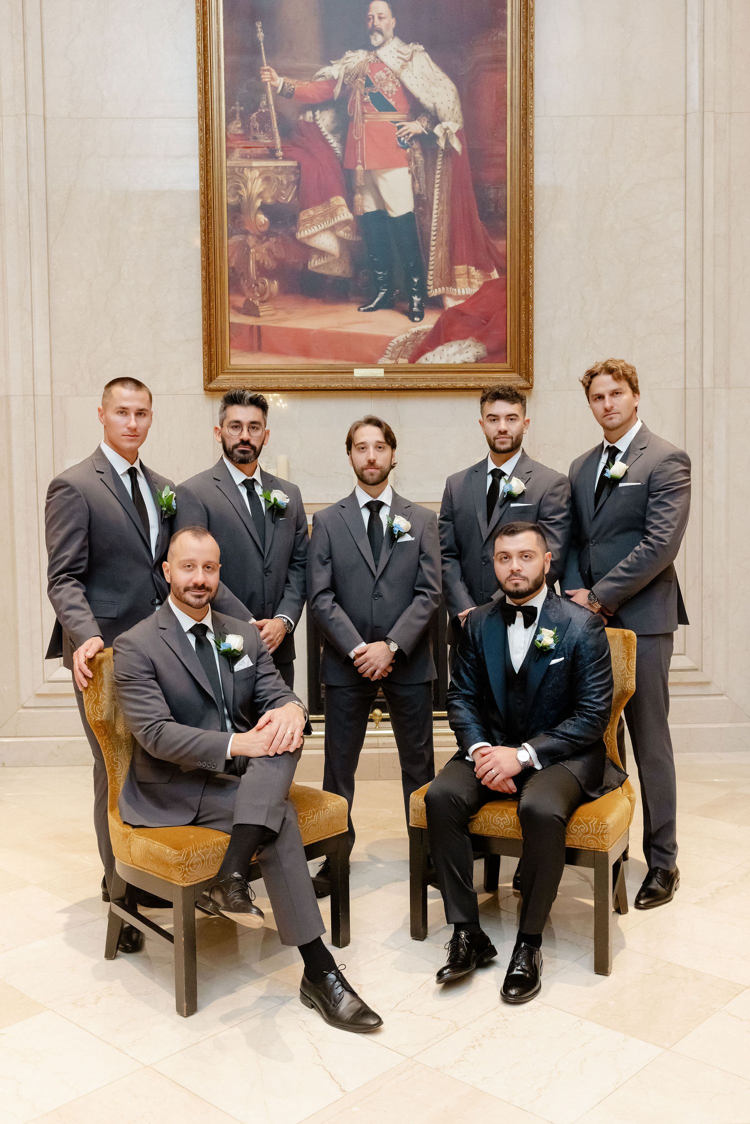 Groomsmen in black suits and ties pose together on elegant chairs in a formal setting with ornate framed artwork.