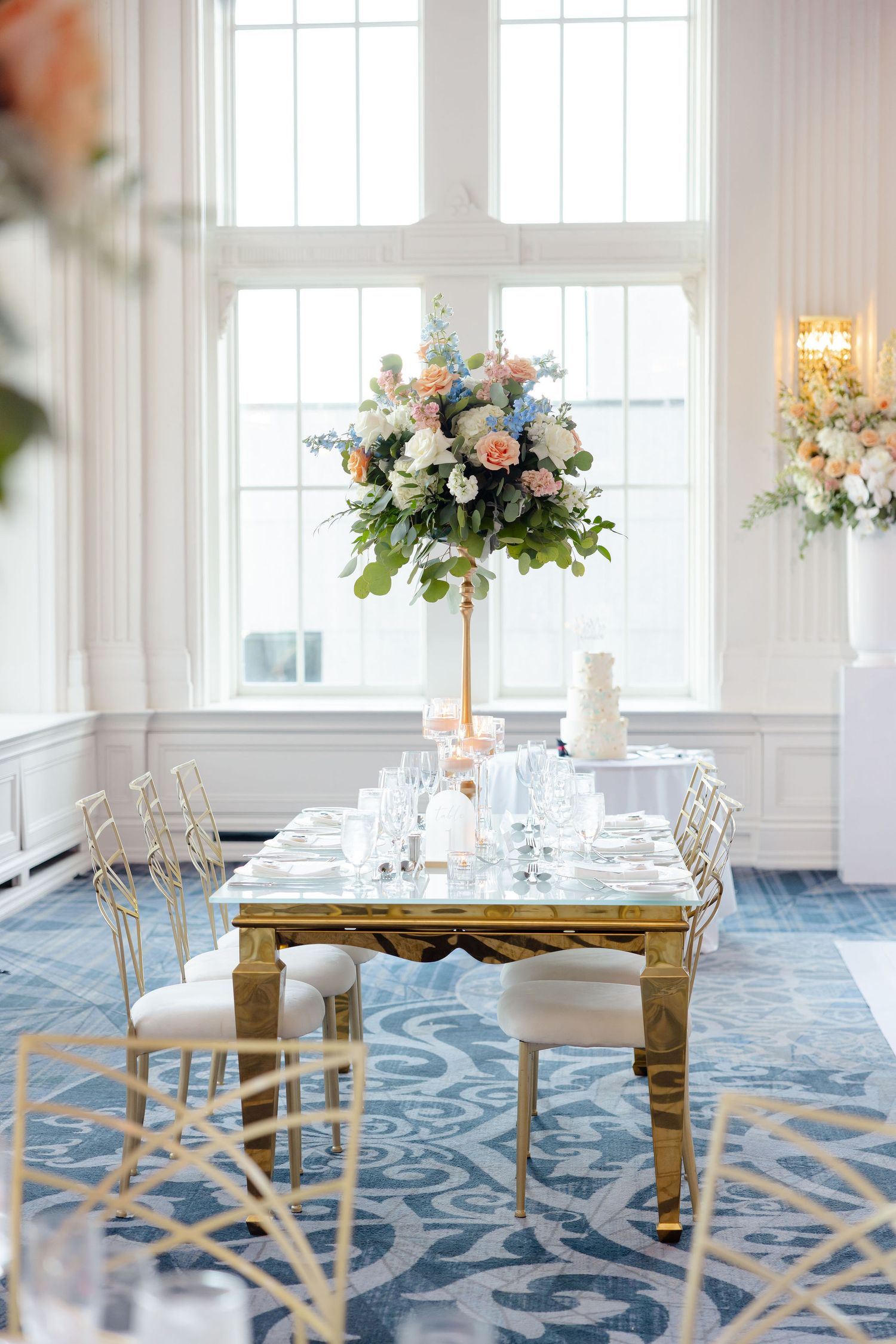 Luxurious wedding reception tables with gold decor and blue patterned carpet in a bright white room.