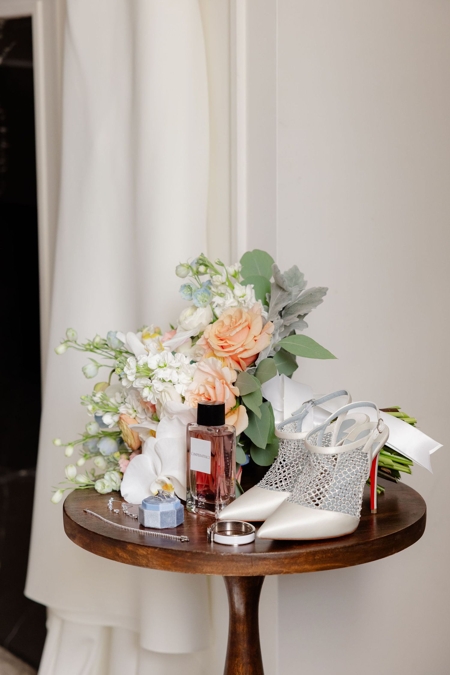 Wedding accessories arranged on wooden table including peach flowers, silver shoes, and perfume bottle.
