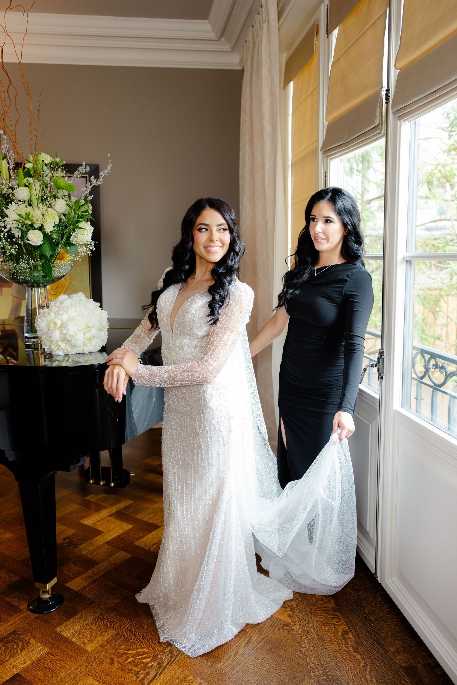 Two women in contrasting white and black dresses pose elegantly by a grand piano and window.