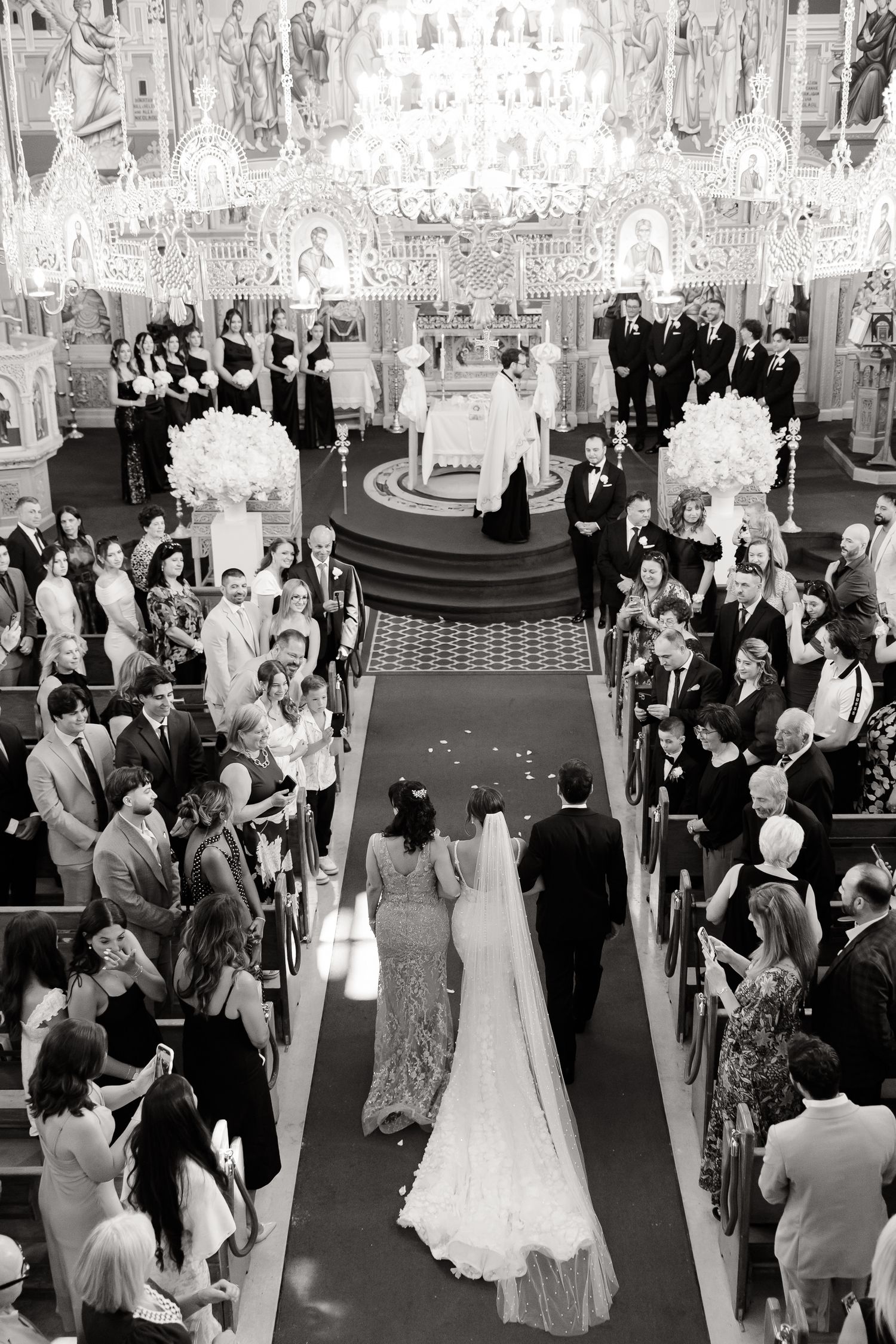 Elegant black and white church wedding ceremony with guests lining the aisle and ornate chandeliers overhead.