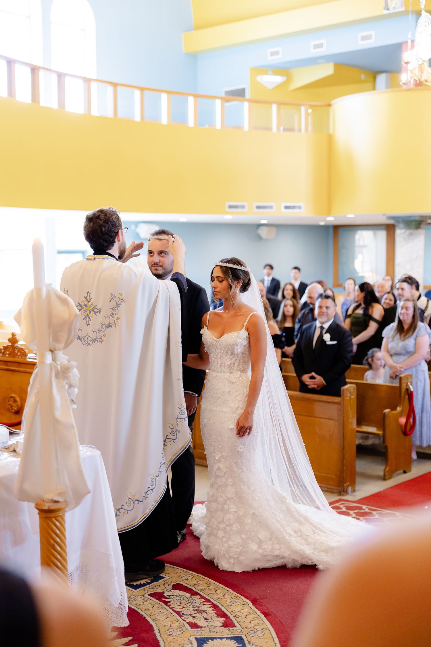 A wedding ceremony takes place in a bright yellow and white church with guests watching from wooden pews.