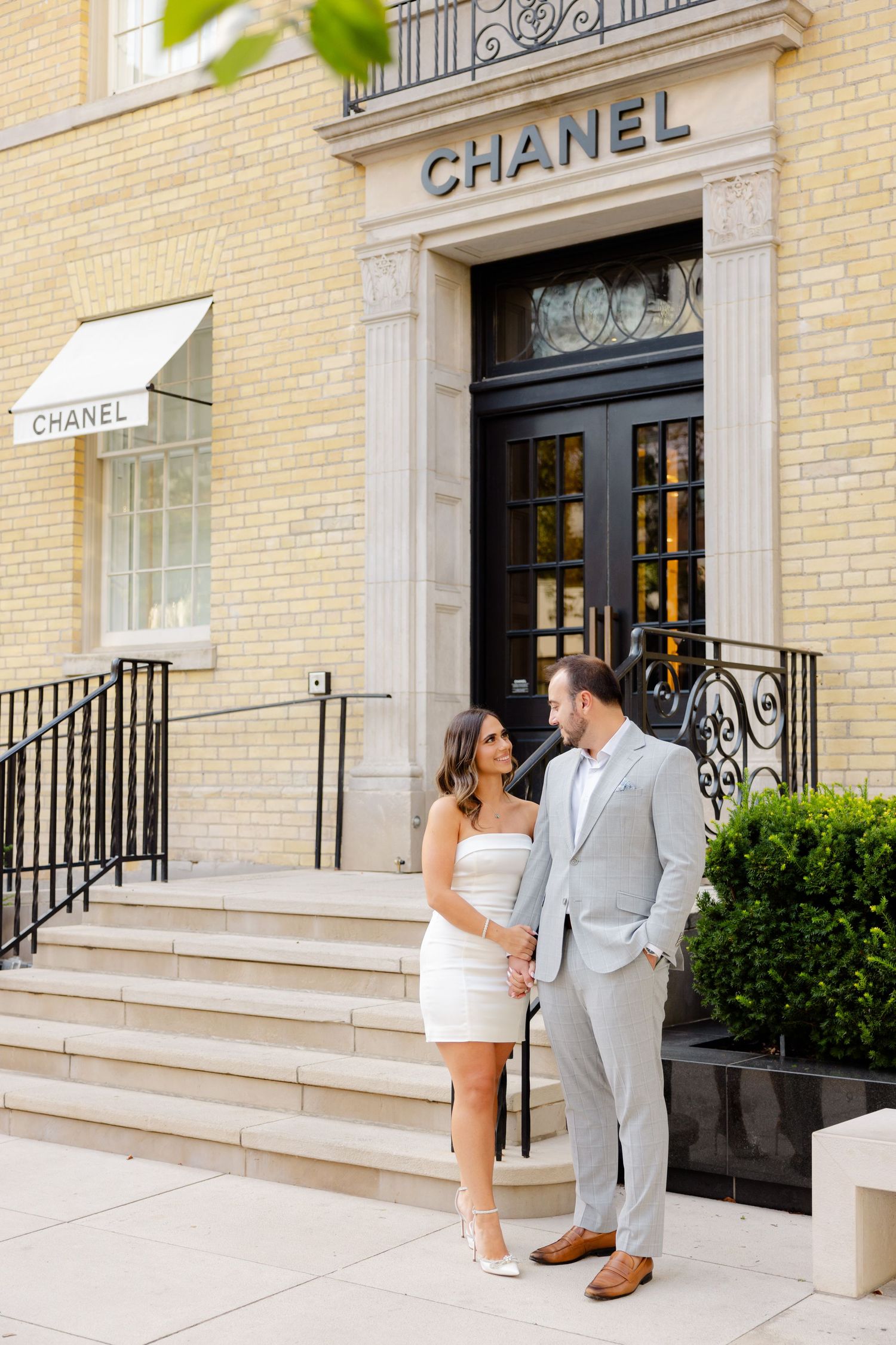 Stylish duo share intimate moment outside Chanel store entrance with ornate architectural details.