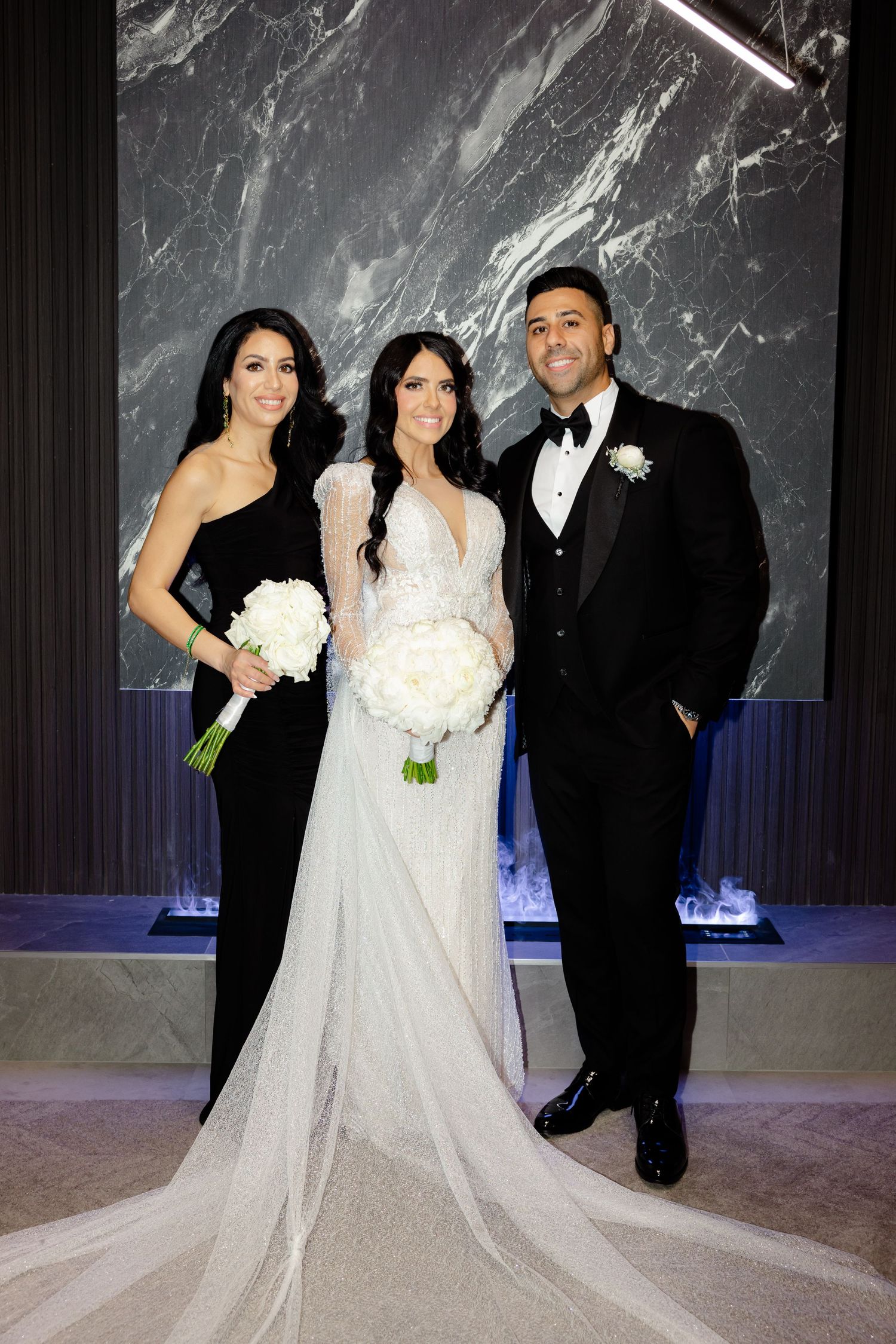 Elegant wedding party portrait with bride in white gown flanked by guests in black formal wear.