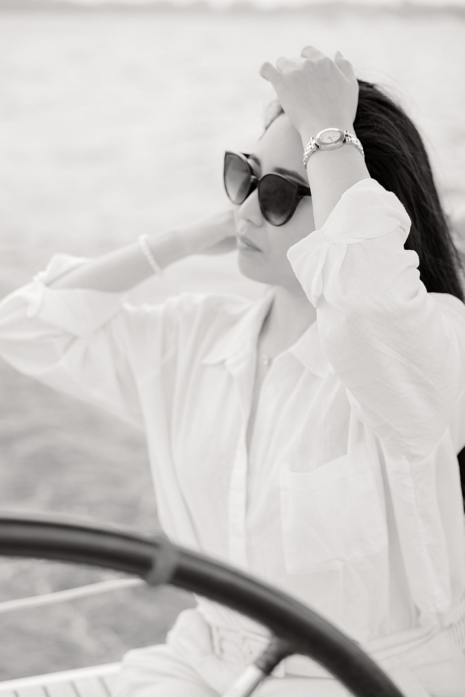 A black and white photo of someone wearing sunglasses and a white shirt, sitting in a car with arm raised to their head.
