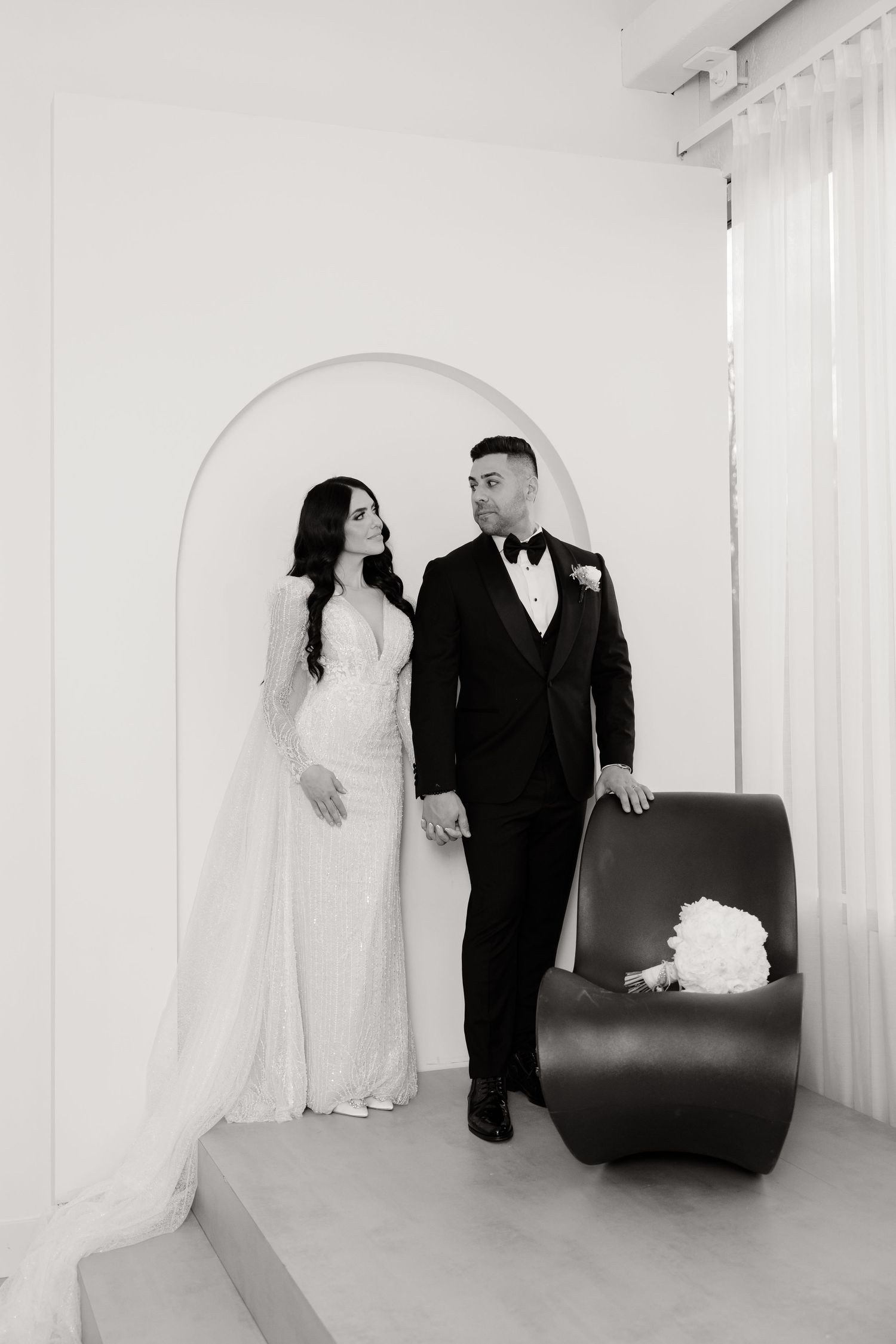 Black and white portrait of newlyweds standing by curved archway with modern furniture.