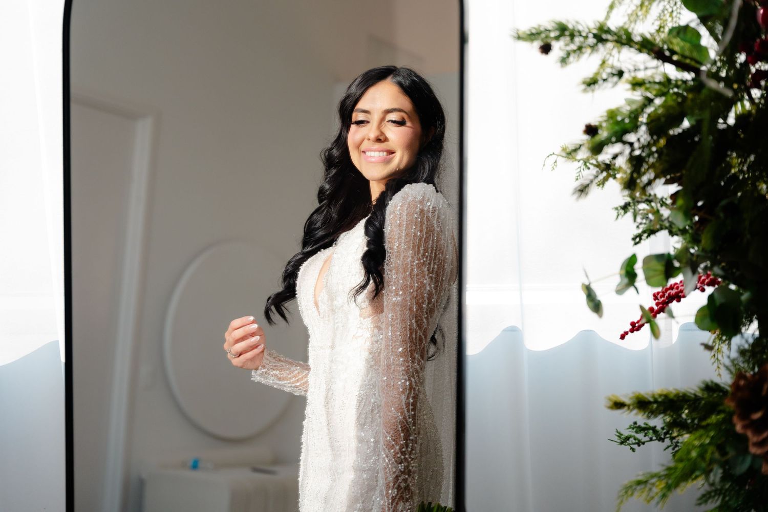 Elegant bride in lace long sleeve wedding dress standing by window with natural light and greenery.