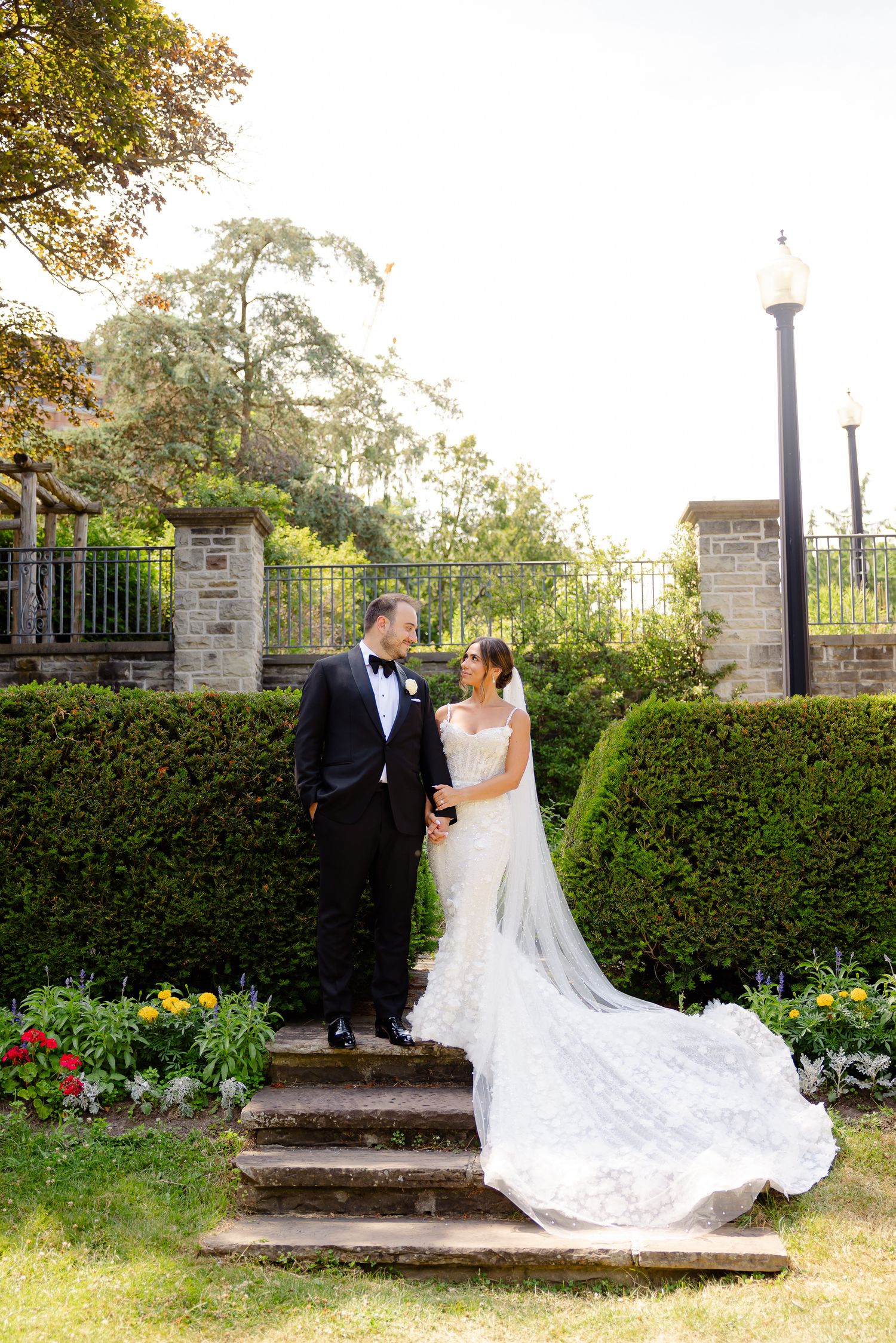 A couple in formal wedding attire pose on garden steps between manicured hedges and ornate lampposts at sunset.