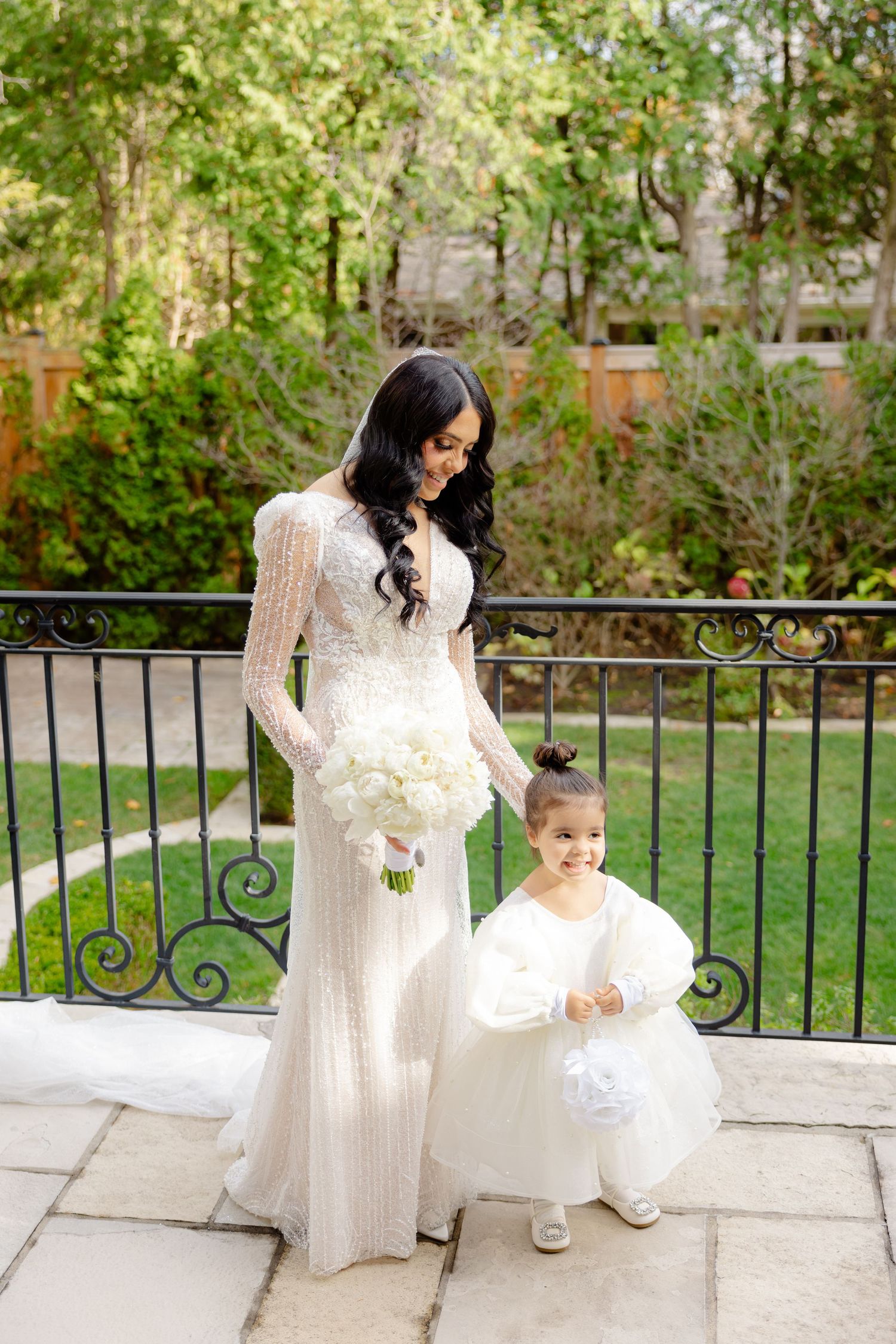 Bride in fitted white dress stands with flower girl in puffy dress holding bouquets on stone balcony.