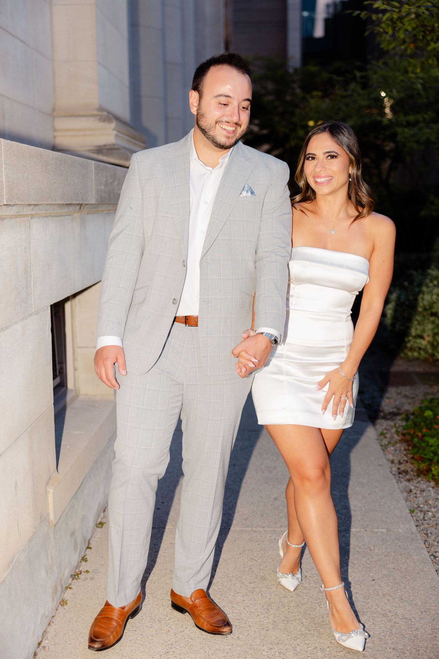 Stylish couple poses together against stone building in evening light.