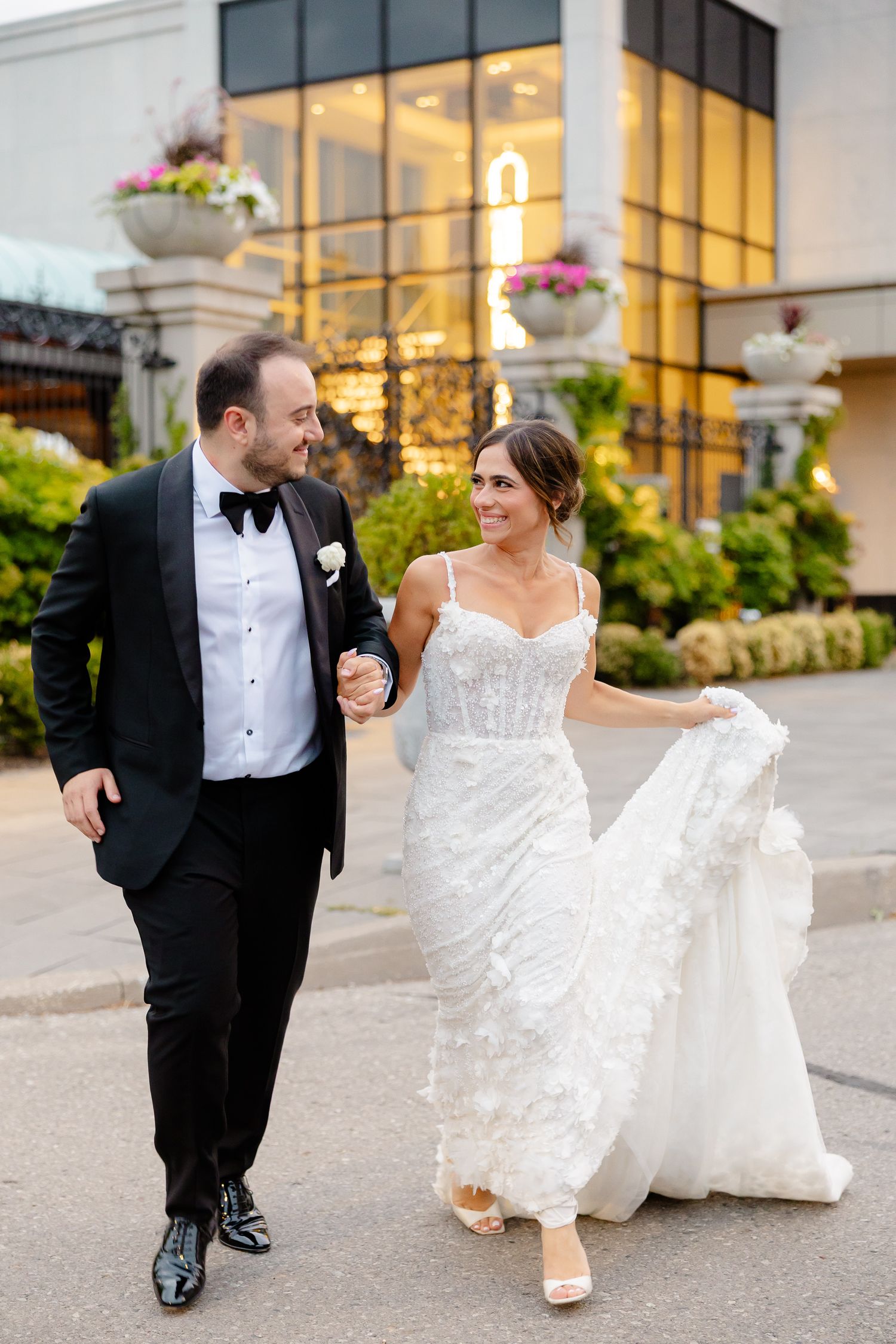 A couple walking outdoors in formal wedding attire with a modern building and hanging flower baskets in the background.