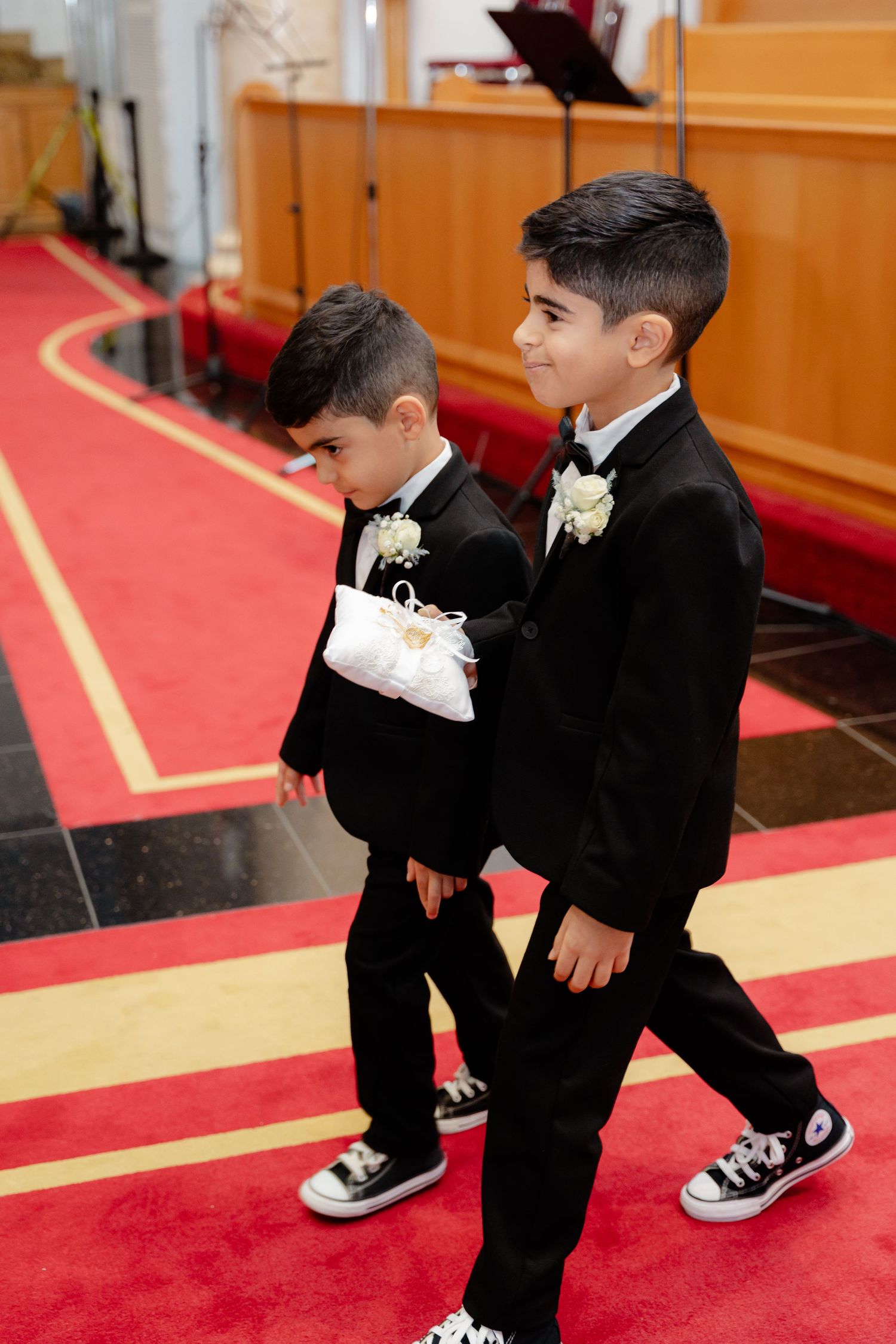 Two young boys in black suits and sneakers walk down church aisle carrying white pillows.