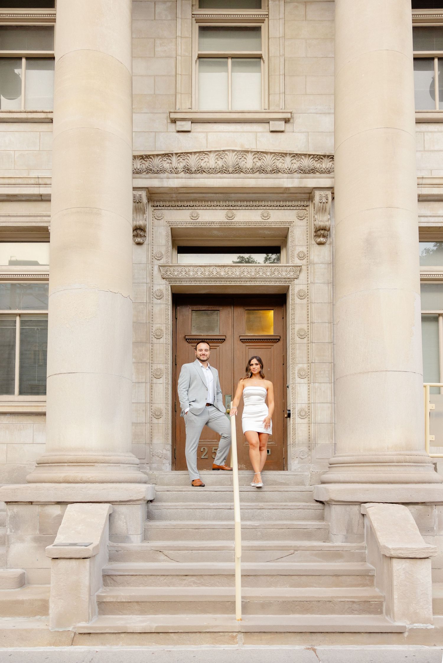 A couple poses in formal attire on grand stone steps of a historic building entrance.
