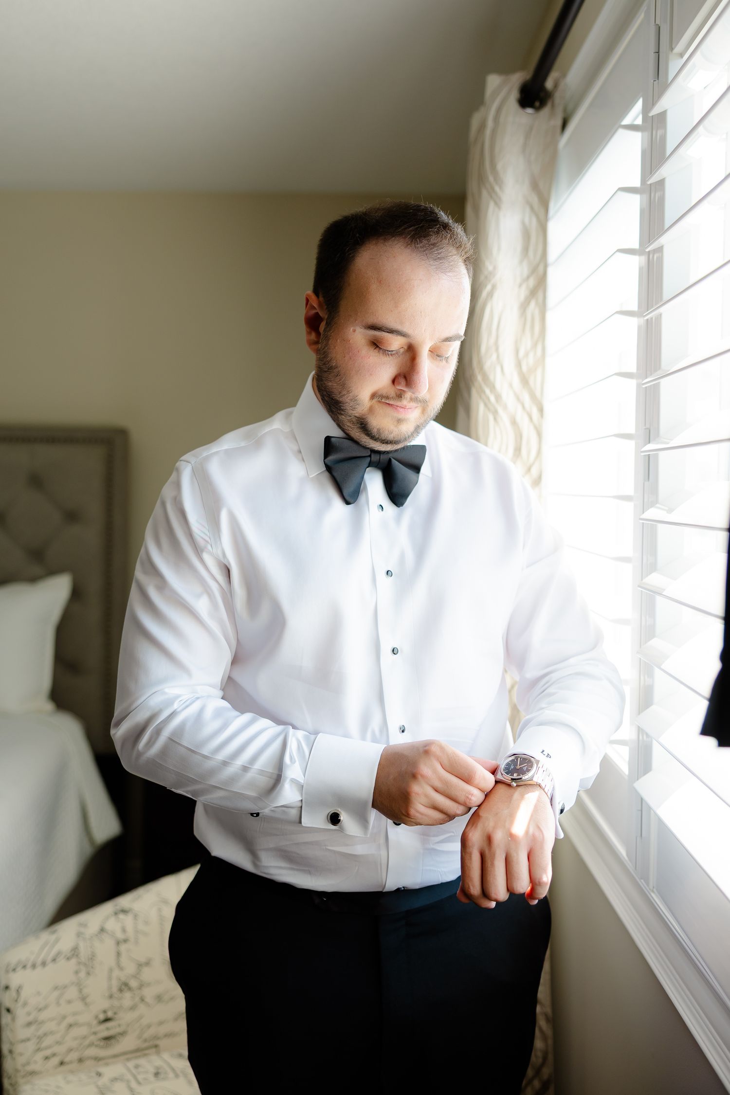Groom getting ready by adjusting his watch near a sunlit window in a white dress shirt.