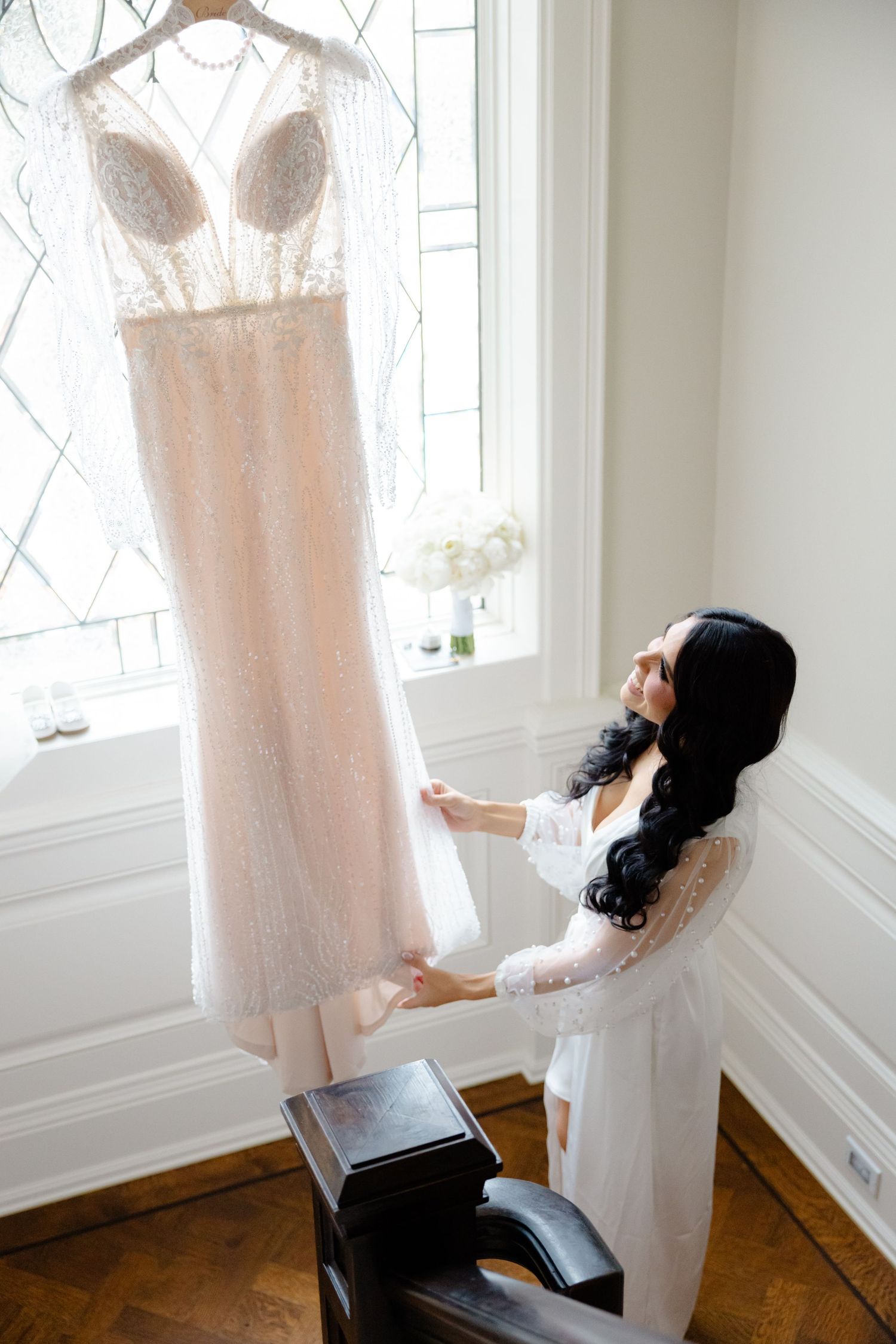 Someone in a white robe admires an elegant blush wedding gown hanging in front of a window.