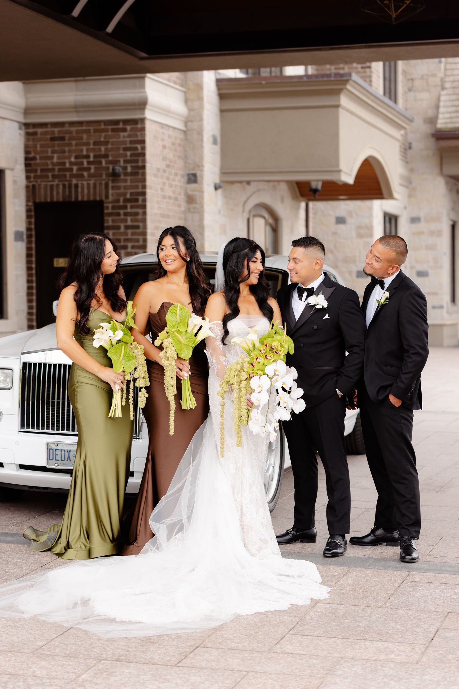 Wedding party poses creatively in front of brick building, bridesmaids in green dresses and groomsmen in black suits.