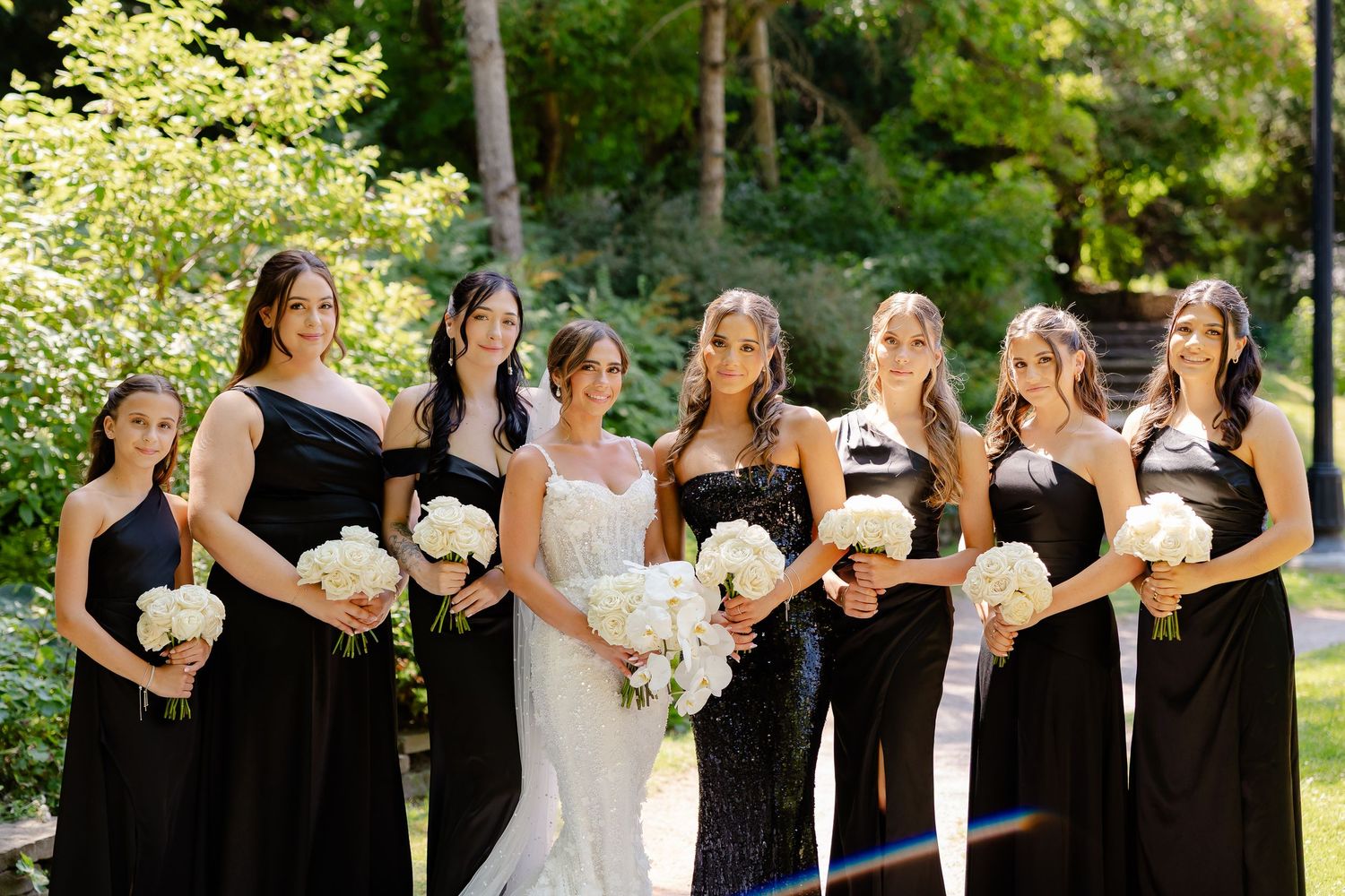 Group wearing elegant black dresses and a white gown pose together with bouquets in a sunny garden setting.