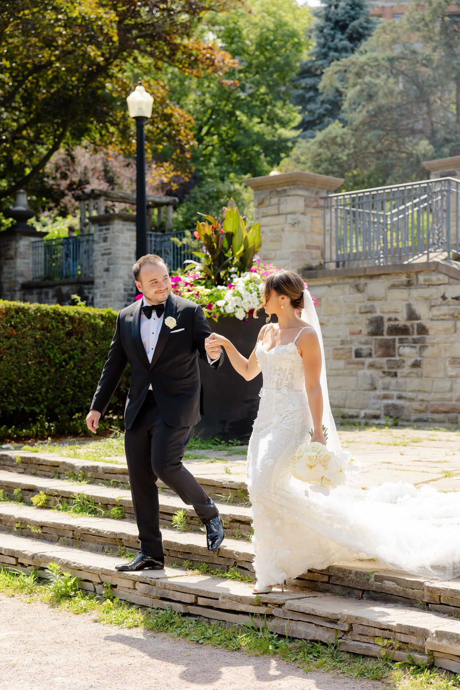 A joyful couple in formal attire descend stone steps on a sunny day surrounded by lush greenery and stonework.