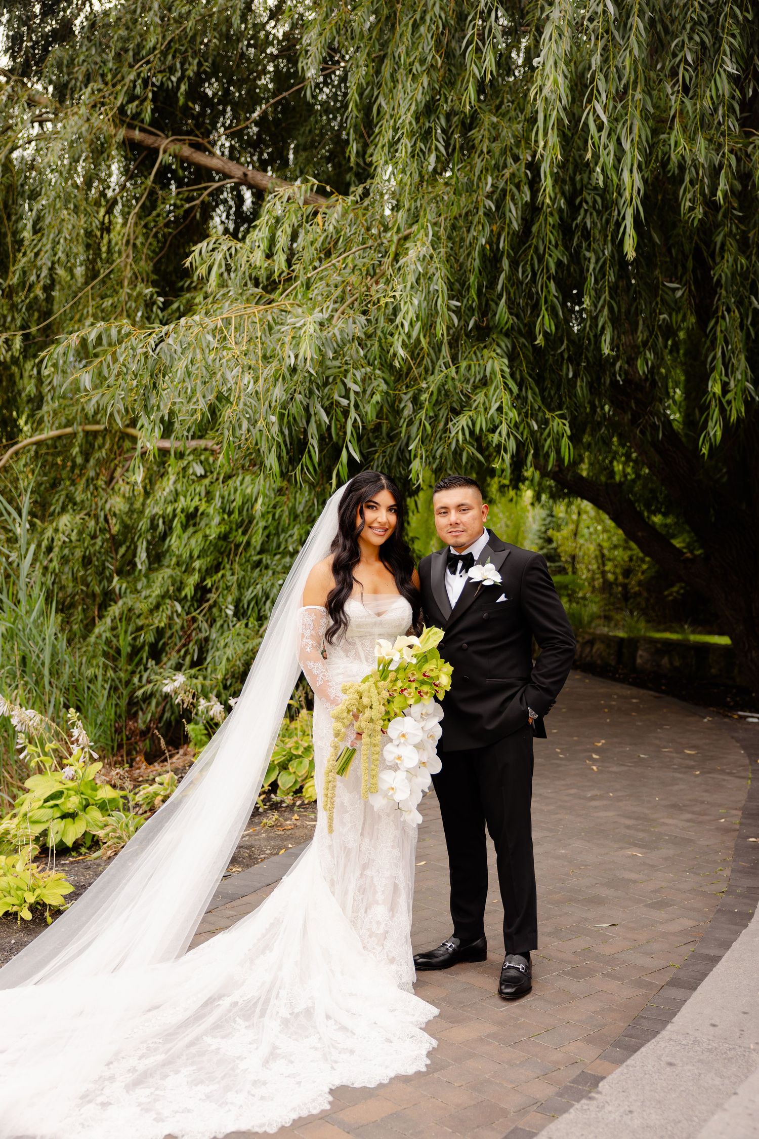 A romantic wedding couple poses elegantly under drooping willow trees on a stone pathway.