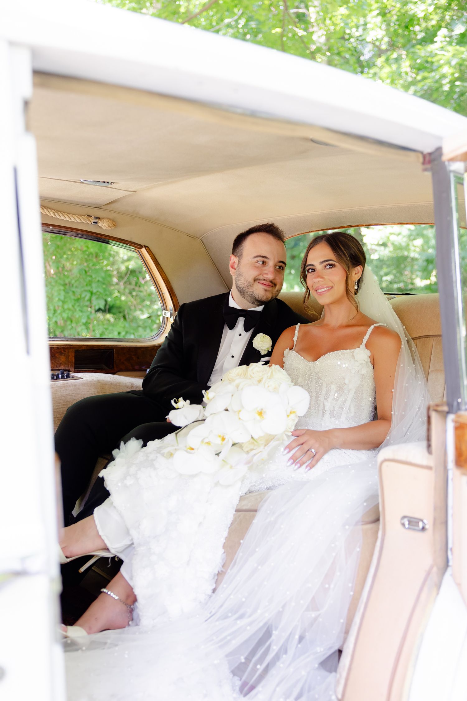 A happy couple sits in a vintage cream-colored car on their wedding day with white floral decorations.