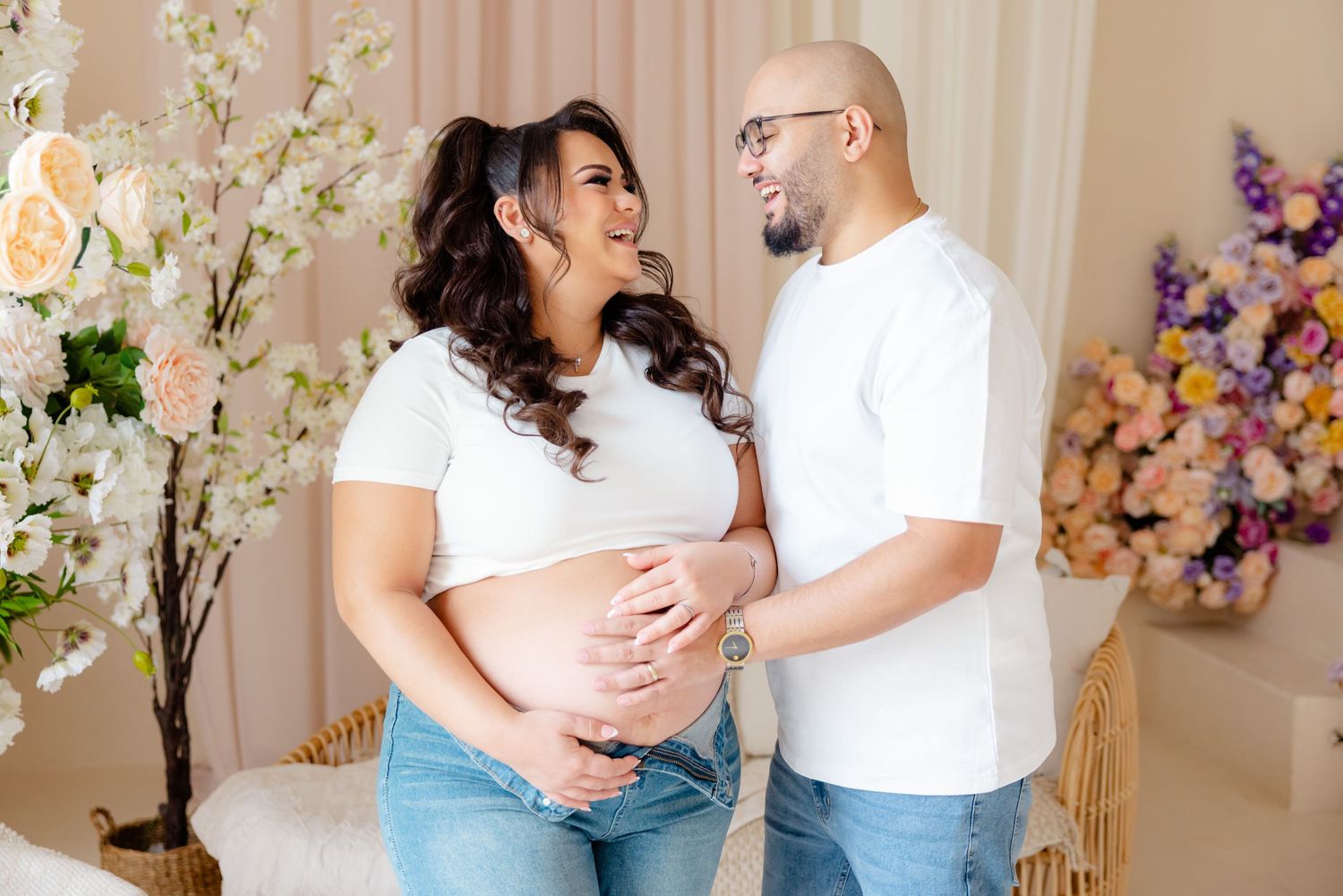 A couple in white shirts share a tender moment during their maternity photoshoot with floral decorations.