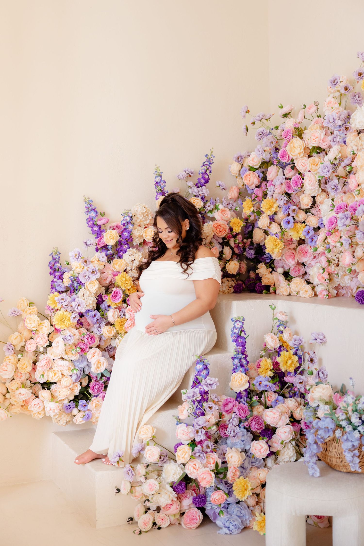 Indoor maternity photo with white dress against wall decorated with pink, purple and yellow floral arrangements.