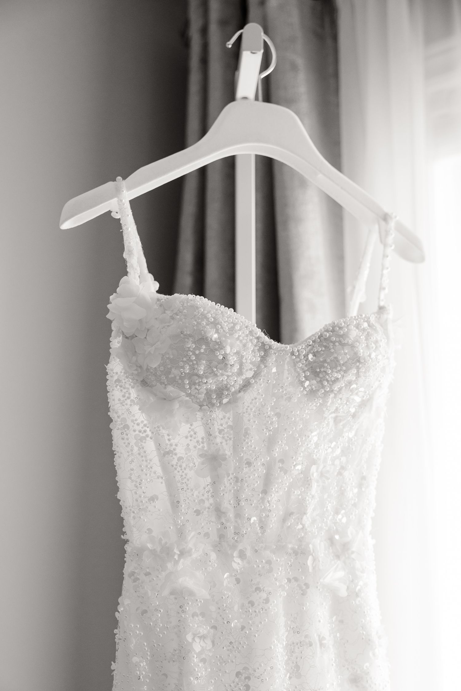 Several elegant white wedding dresses with delicate beading details hang from satin hangers in a boutique setting.