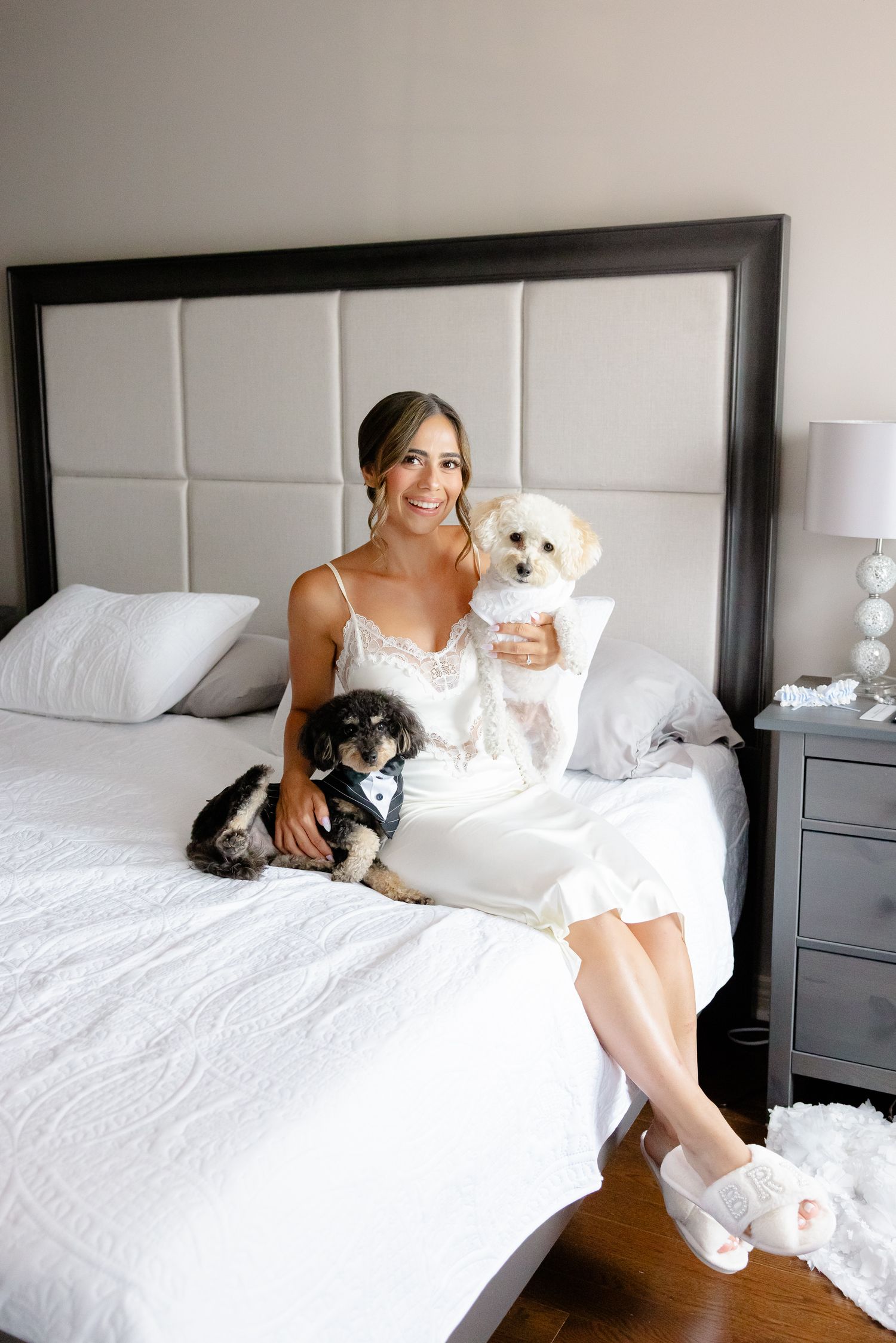 Woman in white dress sitting on bed with two small dogs, one black and one white, in modern bedroom setting.