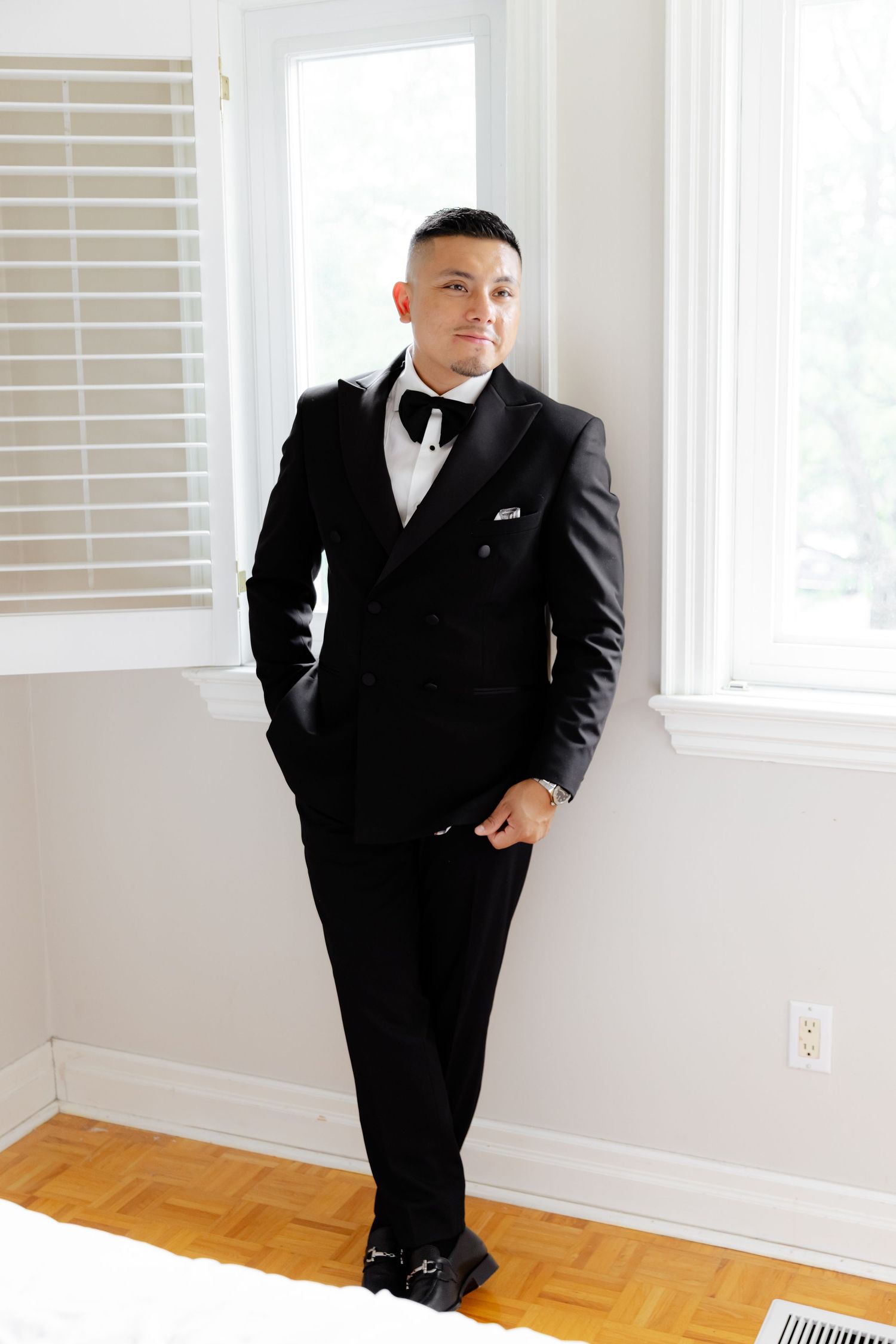 Four images showing someone in a formal black tuxedo and bow tie posing against white walls in an interior room.
