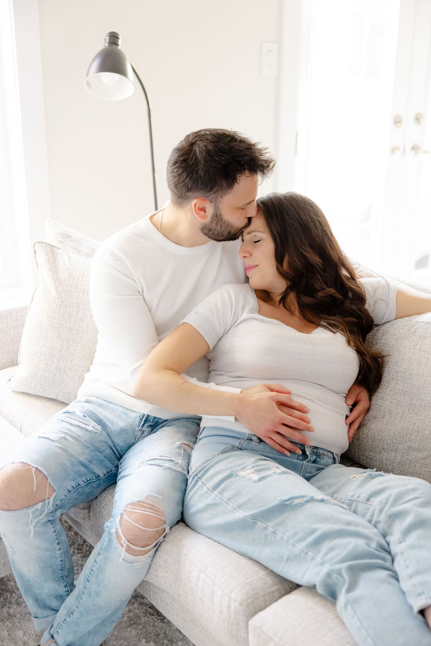Two people embrace on a white couch, with the woman appearing pregnant and wearing light blue jeans.