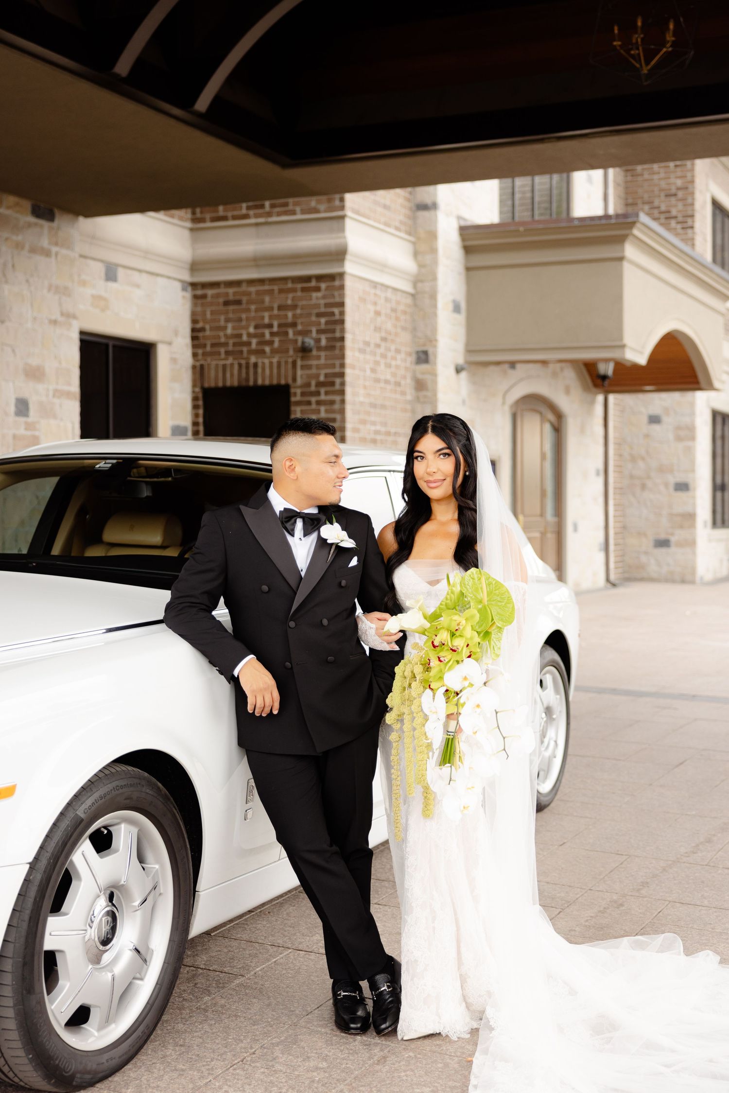 A couple in formal wedding attire poses beside a luxury car parked outside an elegant brick building.