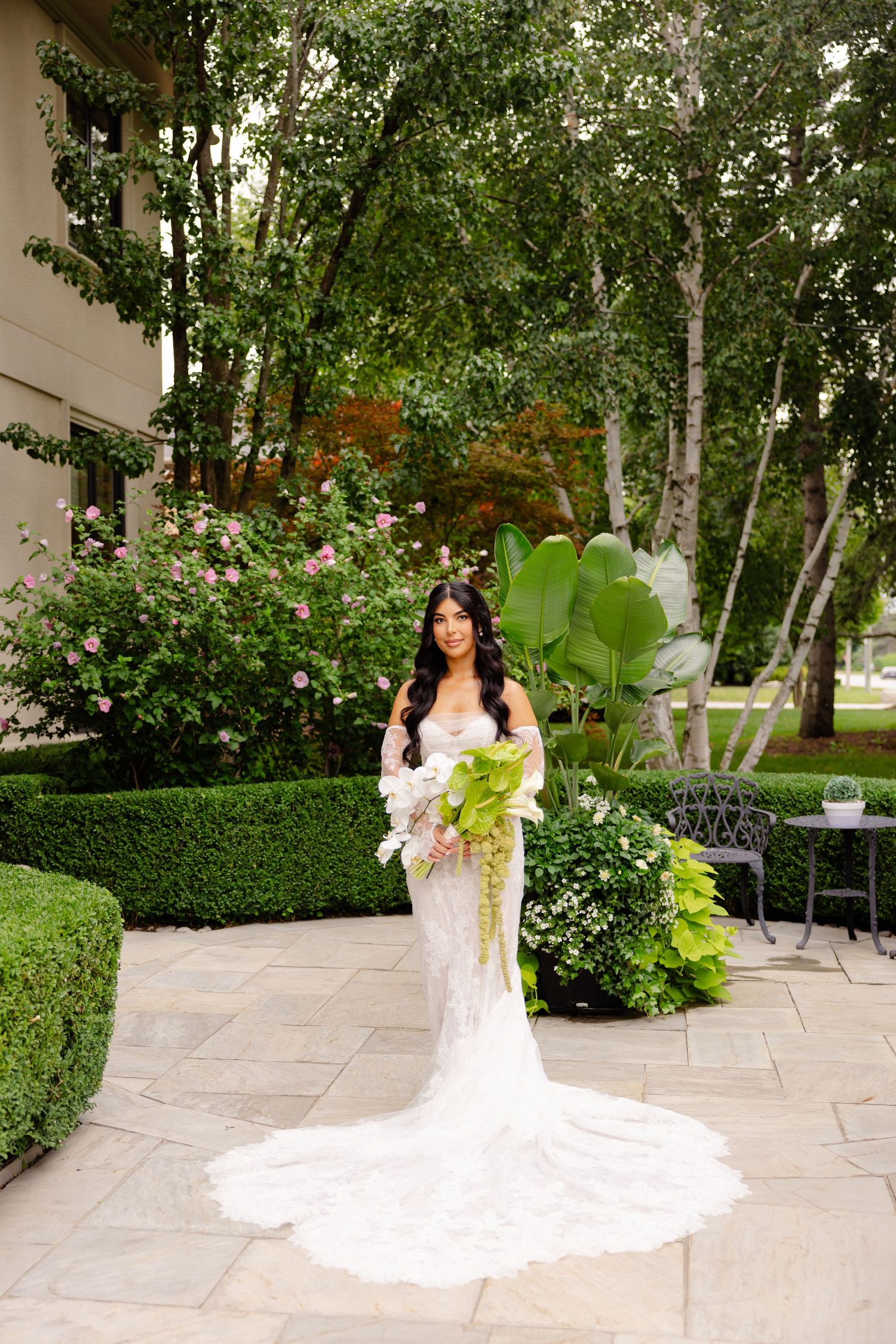 Stunning wedding dress with long train photographed in a garden setting with lush greenery and stone pathways.